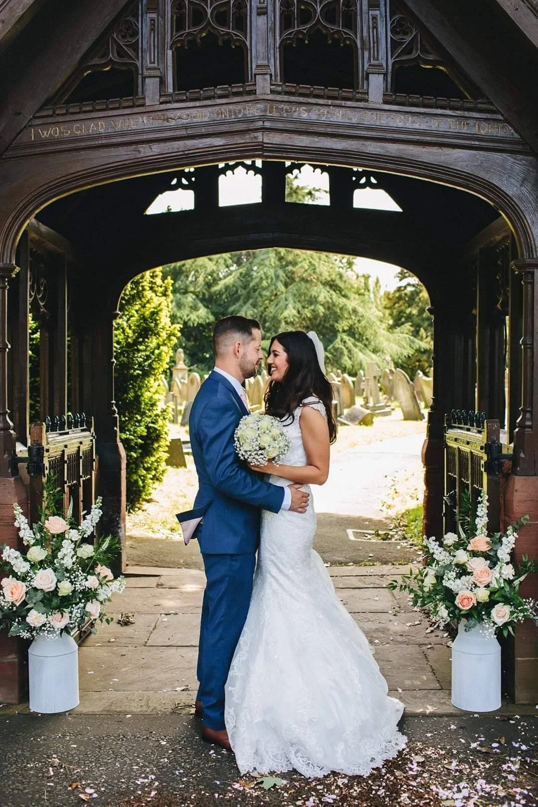 A bride and groom in wedding attire standing close together under a decorative wooden arch in a garden with headstones in the background, surrounded by flower arrangements in white vases.