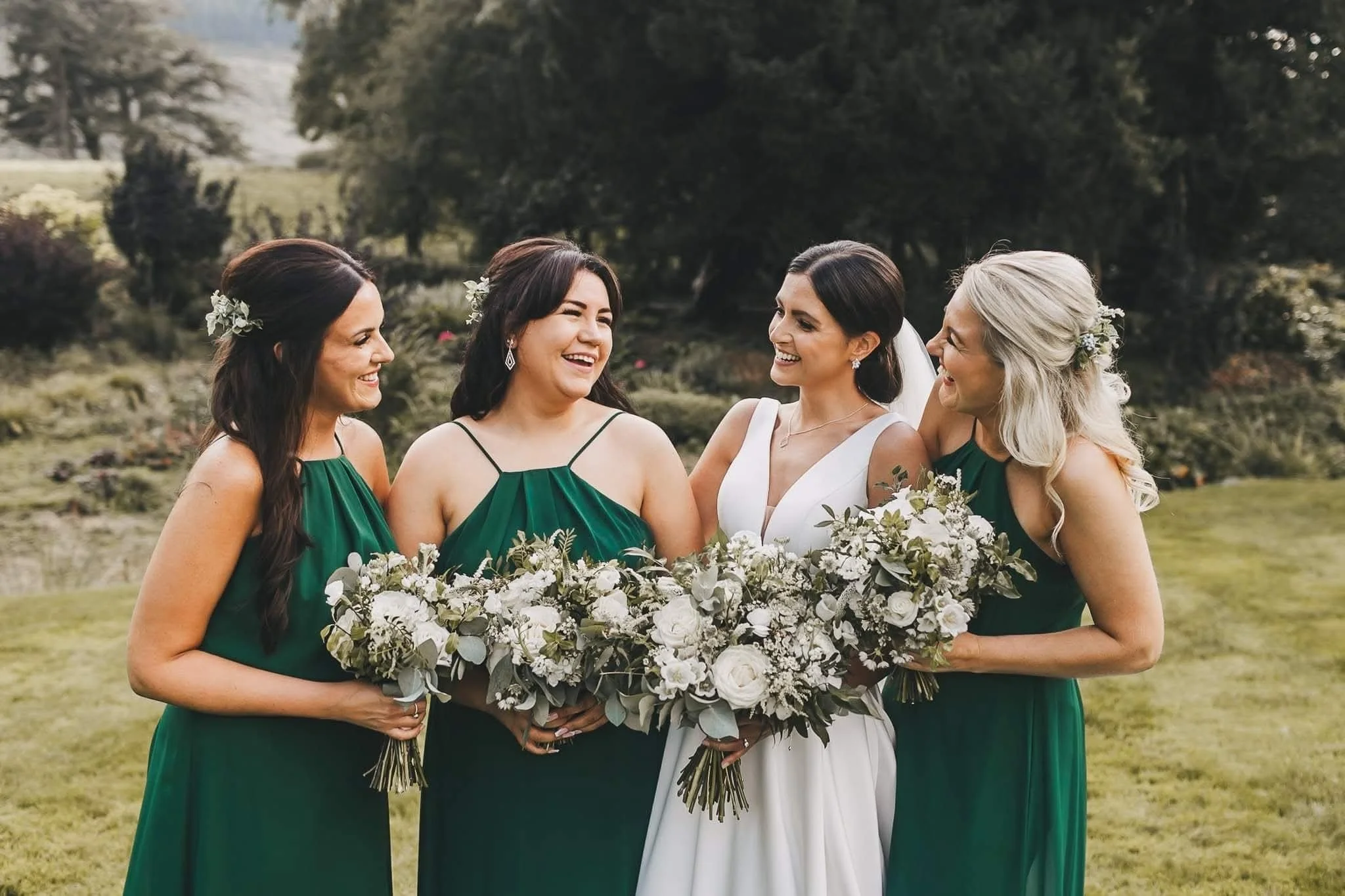 Four women, one bride in a white wedding dress and three bridesmaids in green dresses, standing outdoors on a grassy field with trees in the background, all smiling and holding bouquets of white and green flowers.