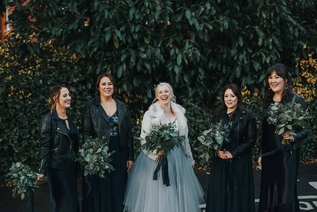 A bride in a light blue wedding dress and a white fur jacket holding a bouquet of green foliage, standing with five bridesmaids dressed in dark clothes and black leather jackets, holding similar bouquets, in front of a large leafy hedge, smiling and 