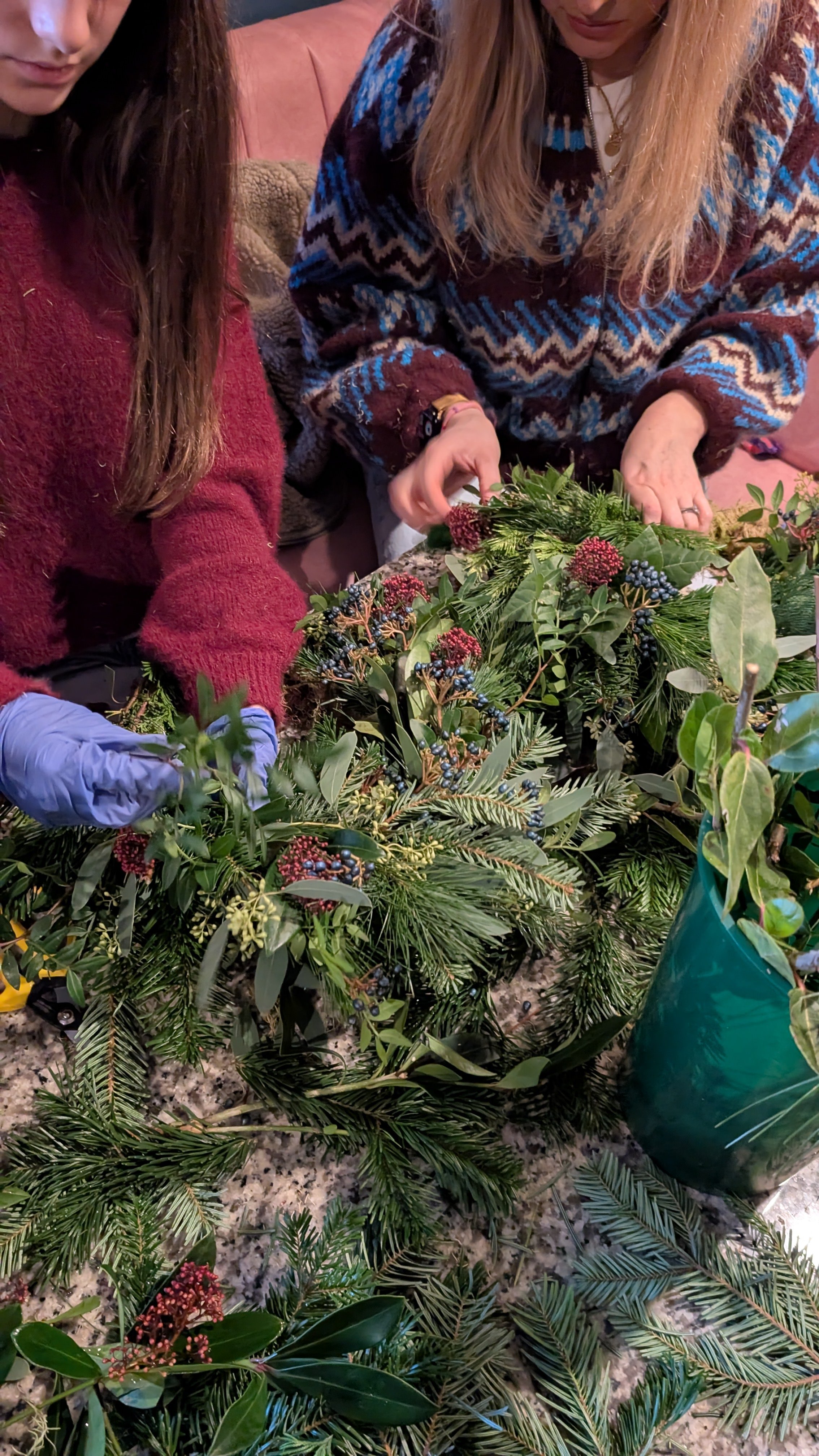Two women wearing sweaters and gloves are arranging Christmas greenery with red and blue berries on a granite countertop, with a green vase holding more greenery nearby.