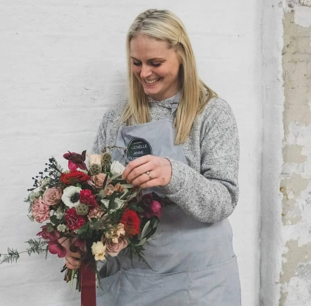 A smiling woman with blonde hair wearing a gray apron standing against a white wall, holding a colorful bouquet of flowers.
