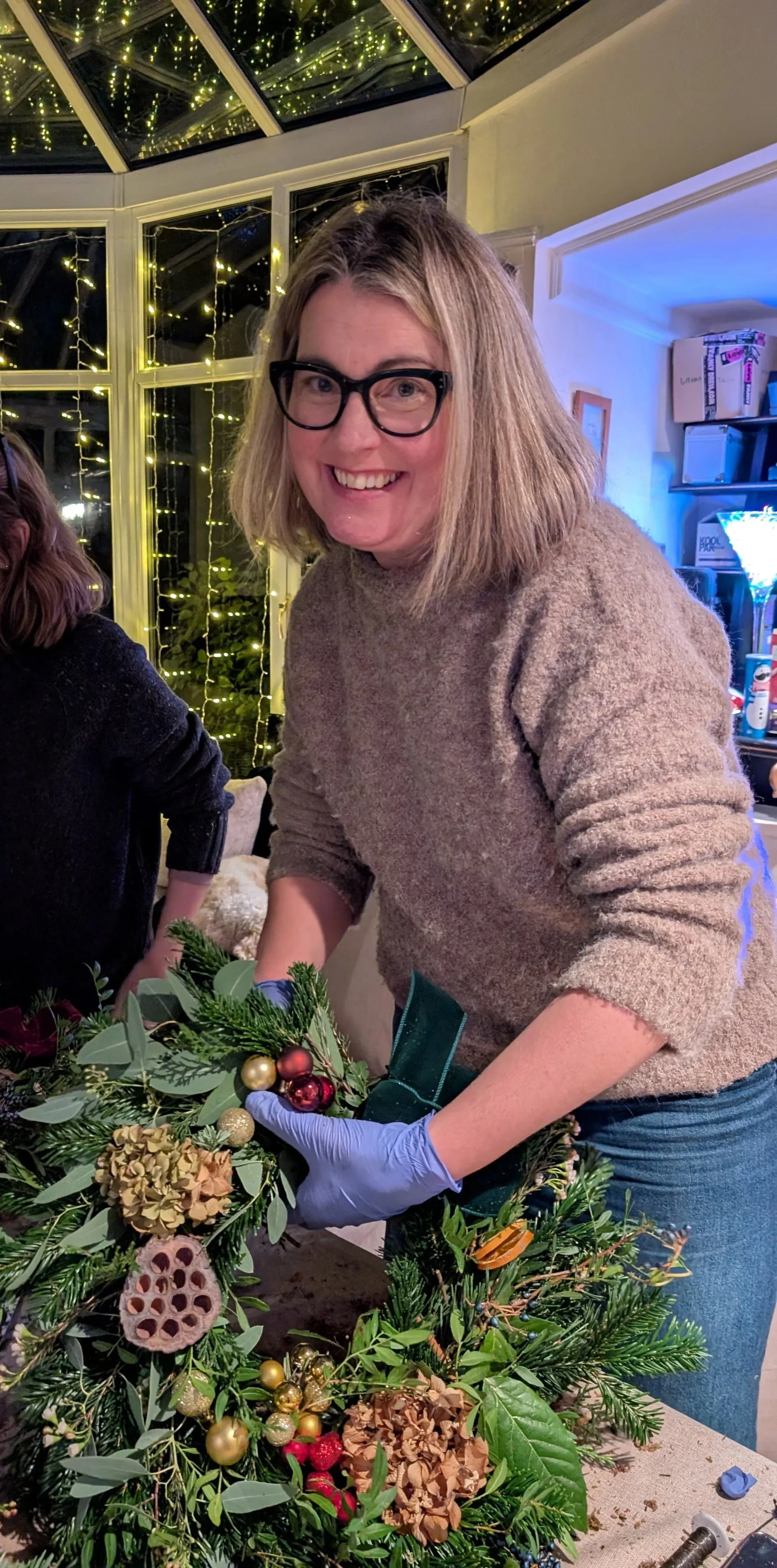 Woman wearing glasses and a brown sweater decorating a Christmas wreath with ornaments in a room decorated with string lights.
