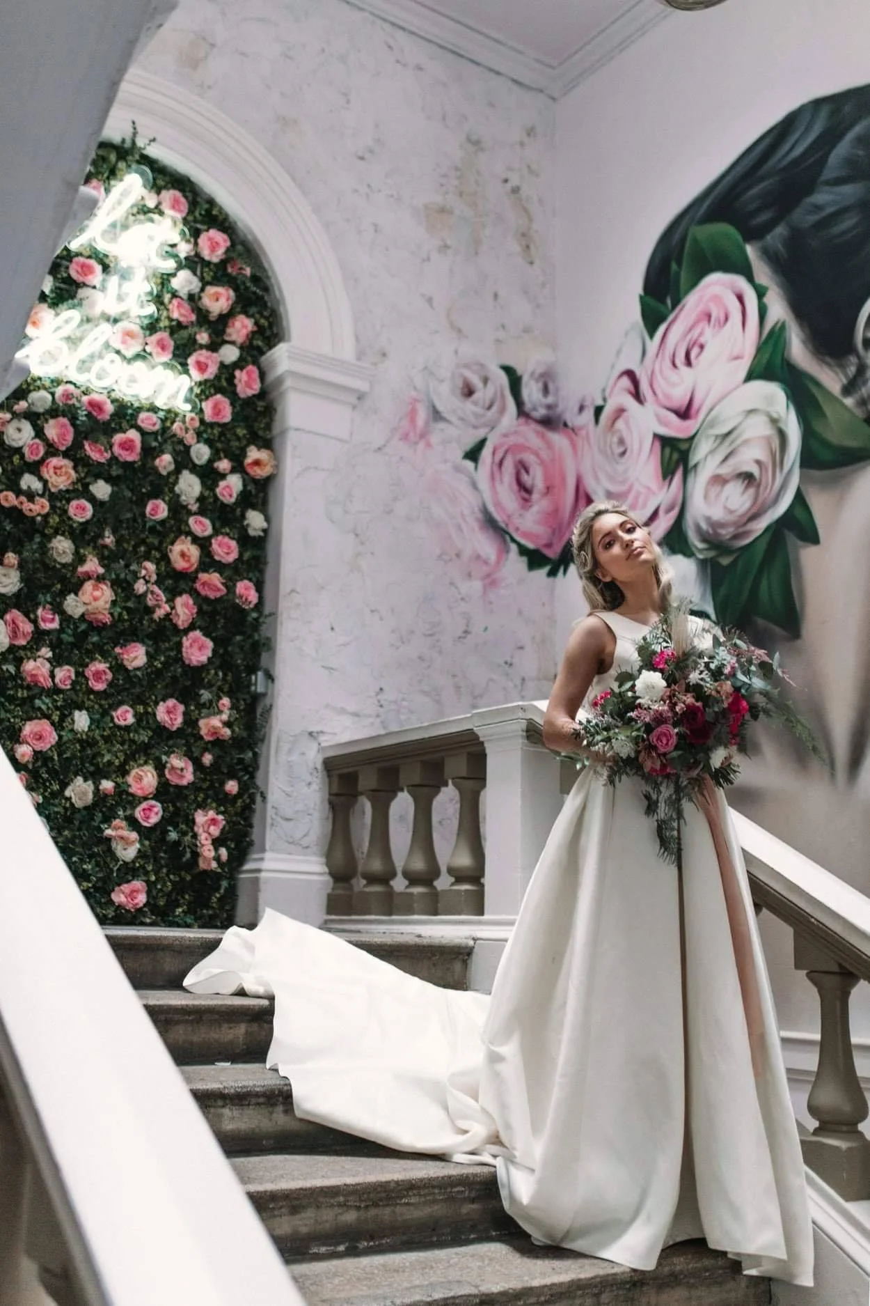 A bride in a white wedding gown holding a bouquet of pink and white flowers, standing on stairs near a large mural of flowers and a neon sign that reads 'Hello Blossom' inside a building with decorated walls.