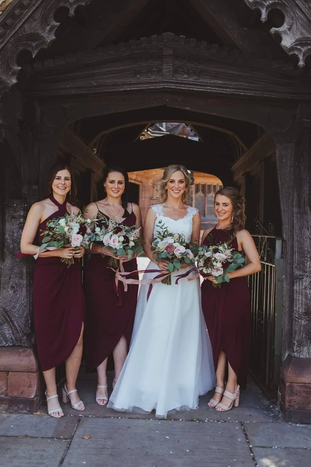 A bride in a white wedding dress with three bridesmaids in burgundy dresses holding bouquets, standing under a wooden archway outside a church.