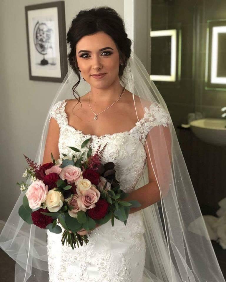 A bride in a white lace wedding dress with a sheer veil, holding a bouquet of pink, cream, and red roses, standing indoors.