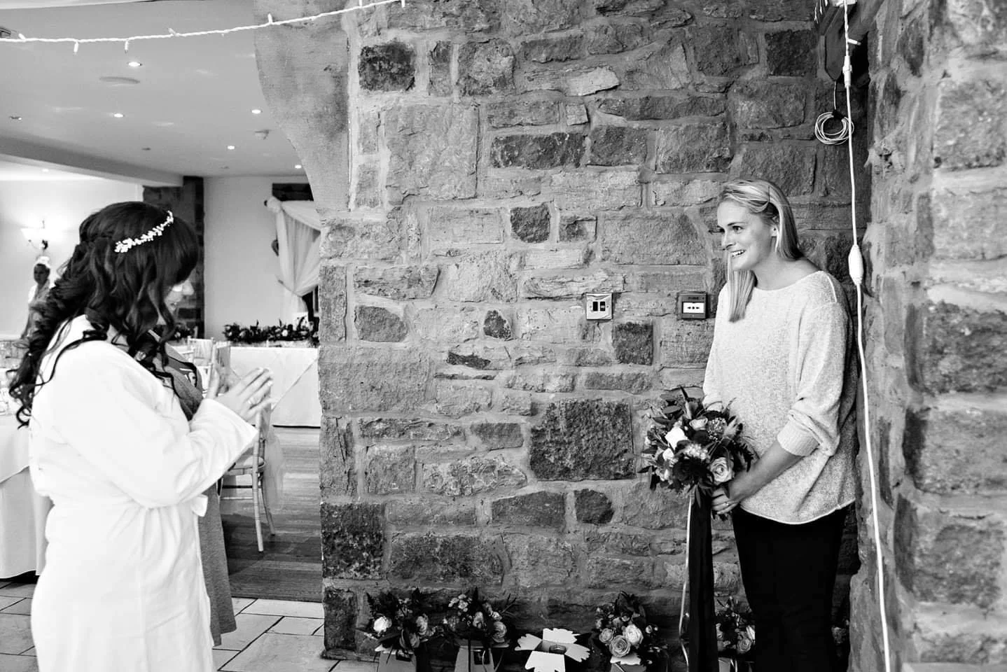 A woman with a bouquet of flowers standing against a brick wall, smiling, while another woman facing her, with her hands clasped, appears to be talking or praying, in an indoor setting with tables and other people in the background.