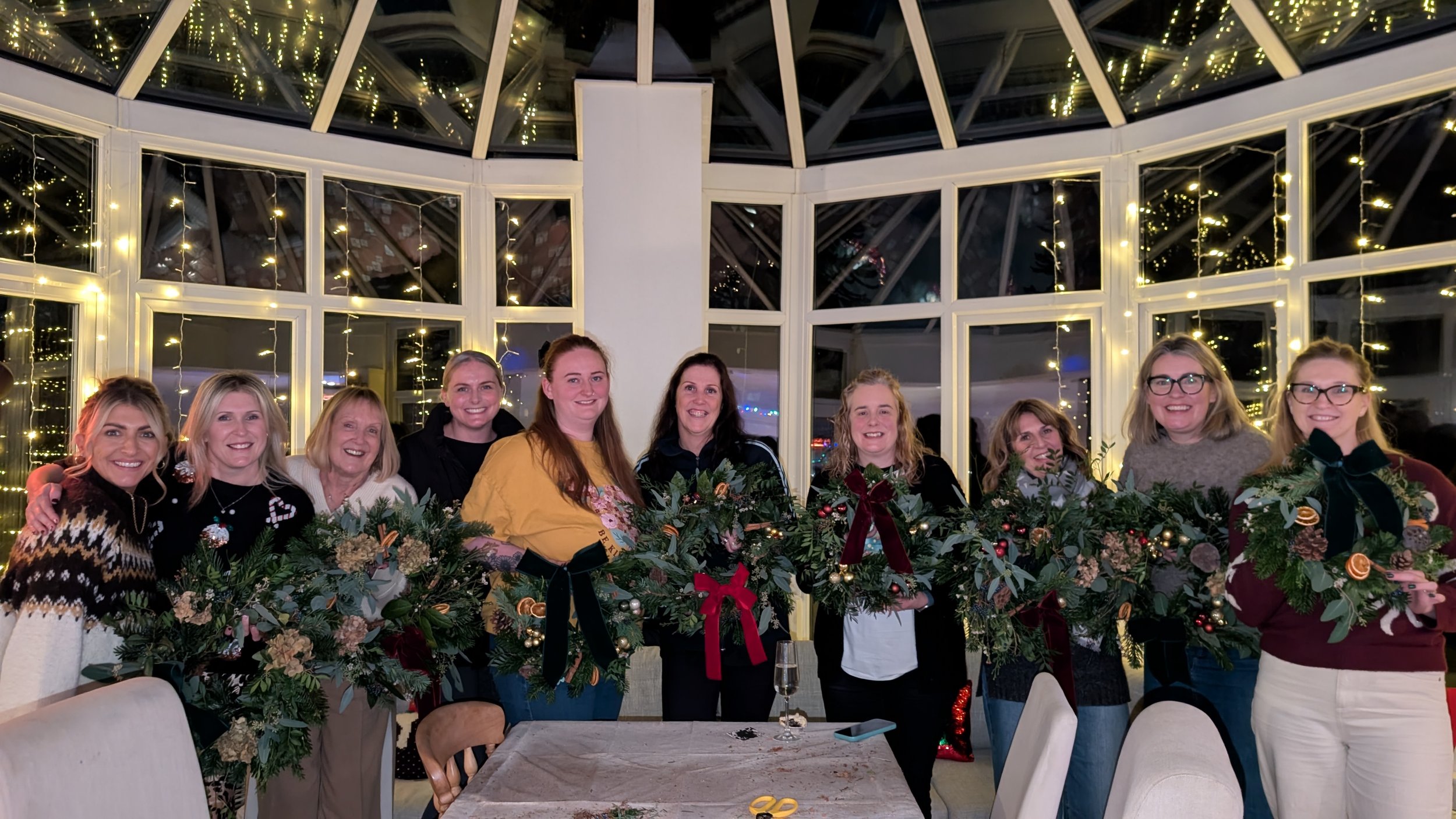 Group of women celebrating Christmas, holding festive wreaths with pine, holly, and ornaments, indoors decorated with string lights.