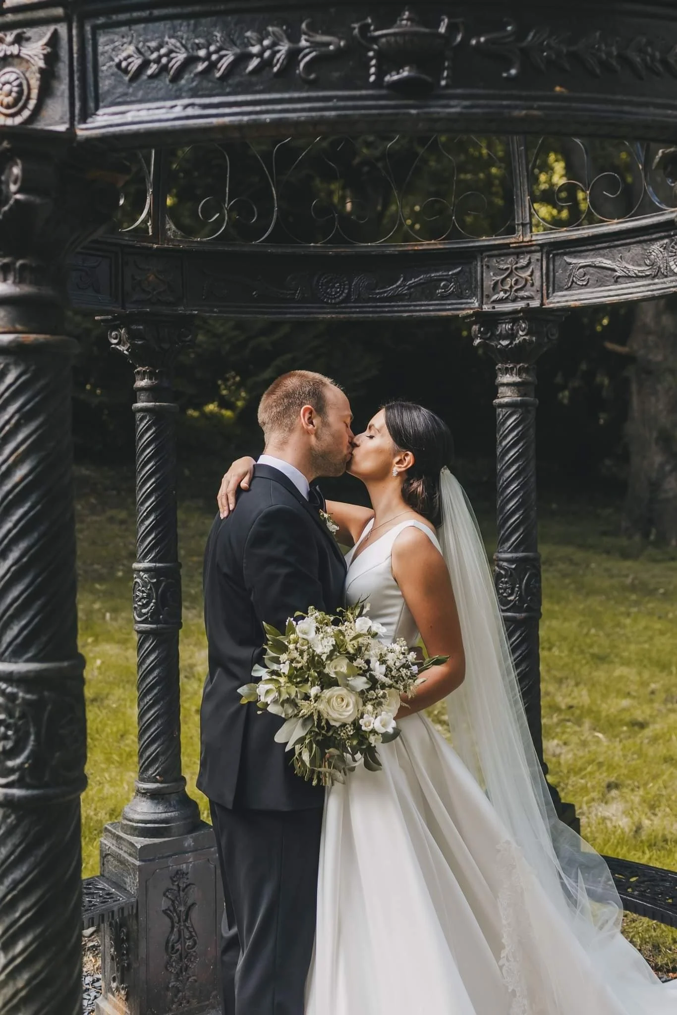 Bride and groom kissing outdoors under a decorative iron structure, with the bride holding a bouquet of white flowers.