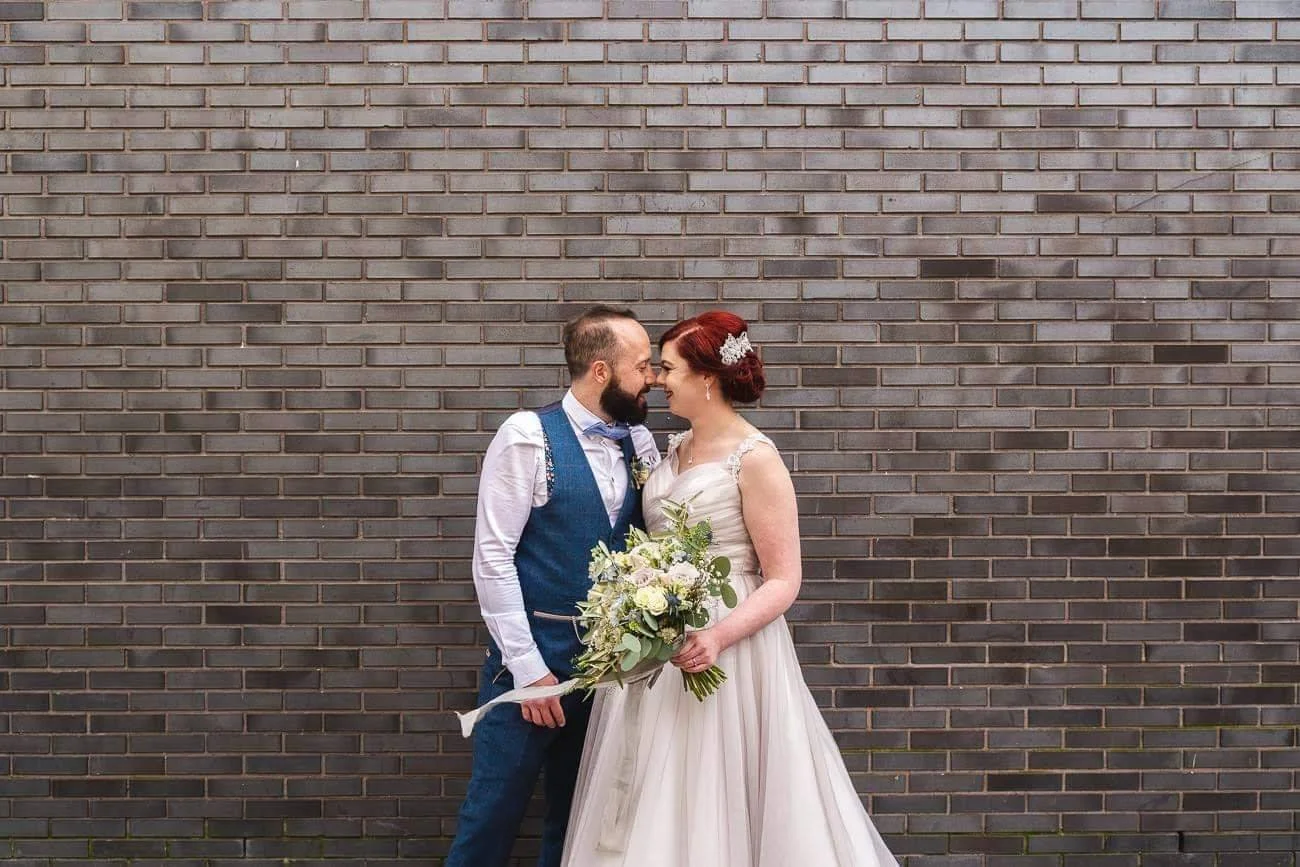 A bride and groom stand close together in front of a brick wall, touching foreheads. The bride holds a bouquet of white and green flowers, and the groom is dressed in a blue vest and pants with a white shirt.