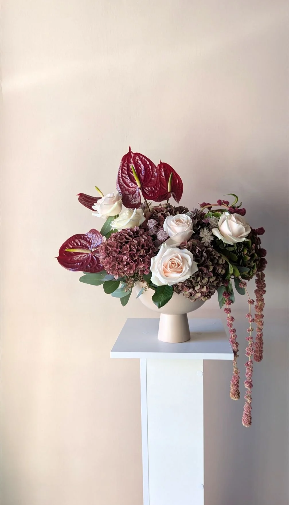 A floral arrangement with pink roses, purple hydrangeas, red anthuriums, and hanging pink amaranthus in a white vase on a white pedestal against a plain wall.