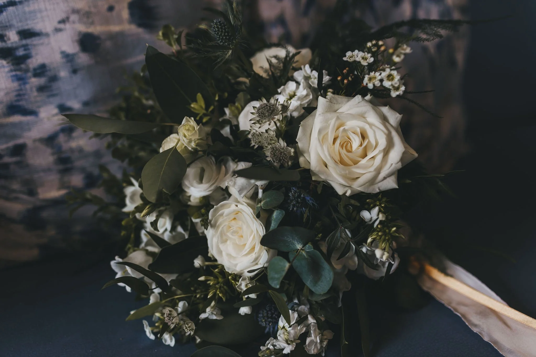A bouquet of white roses, small white flowers, green leaves, and foliage on a dark surface with a blurred background.