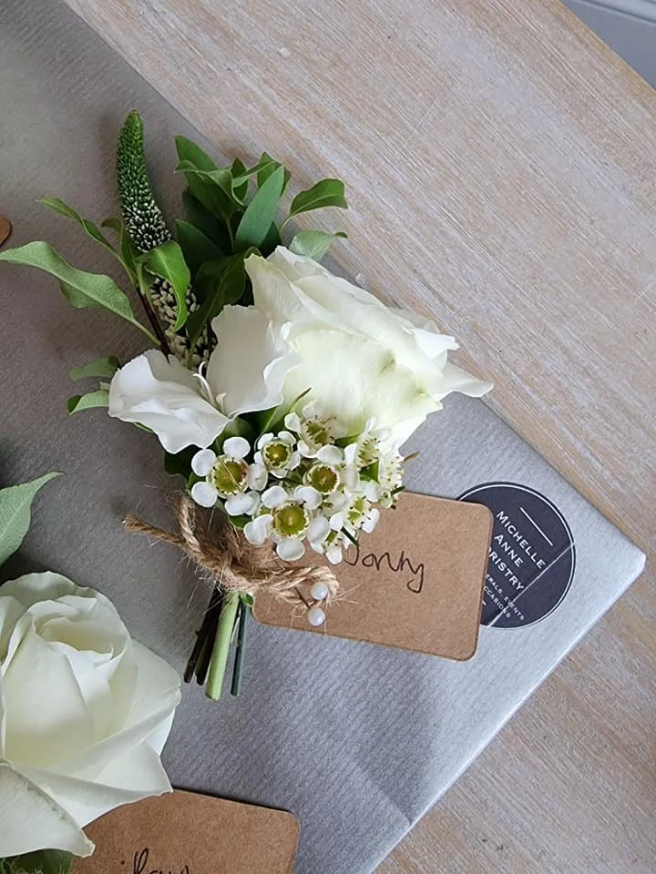 A small bouquet of white flowers, including a white rose and other white blossoms, tied with twine and a brown label with handwriting, resting on a wrapped gift box on a wooden surface.