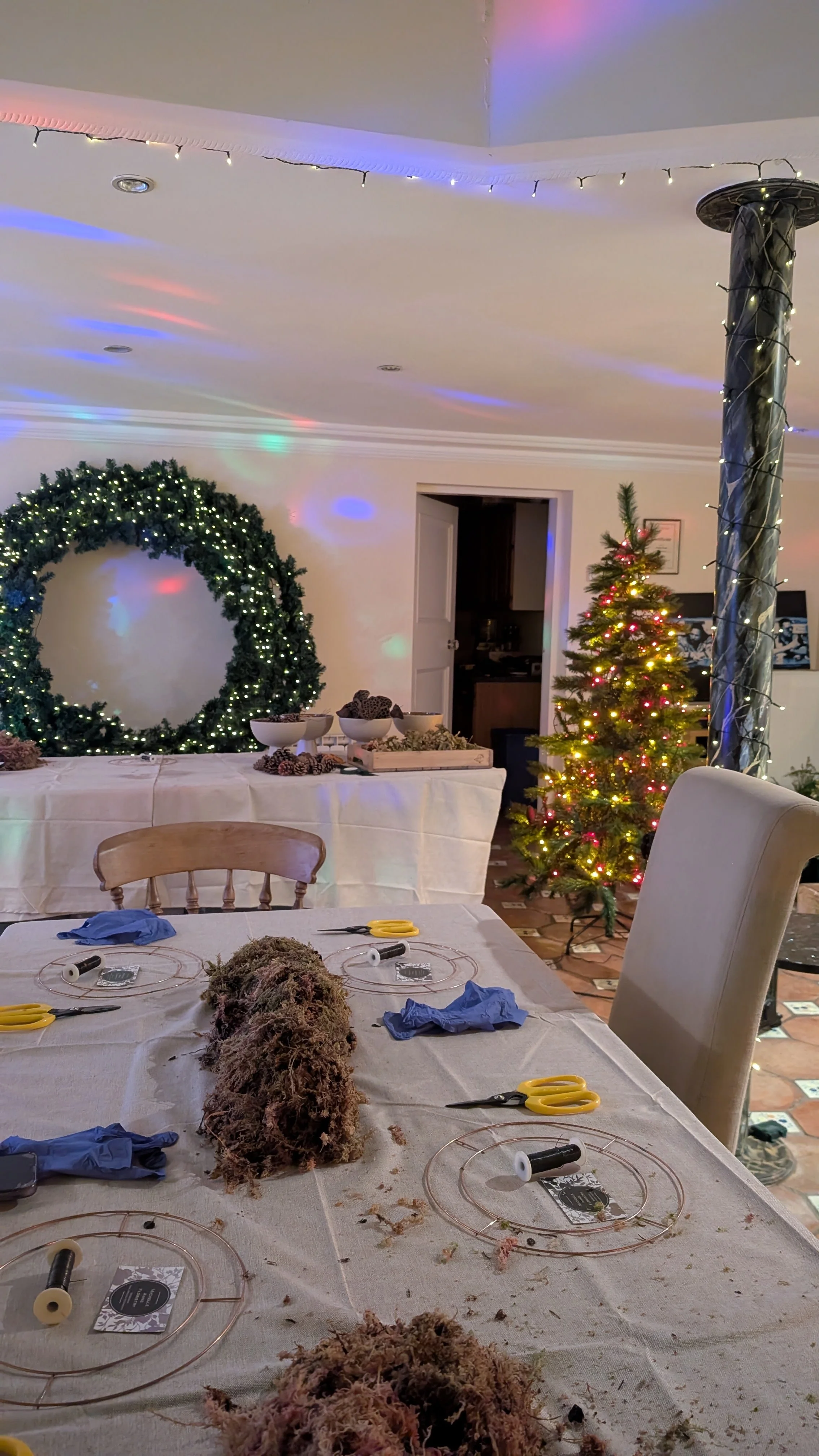 A dining room decorated for Christmas with a small decorated Christmas tree, a large green wreath with lights, and a table with crafting supplies and greenery.