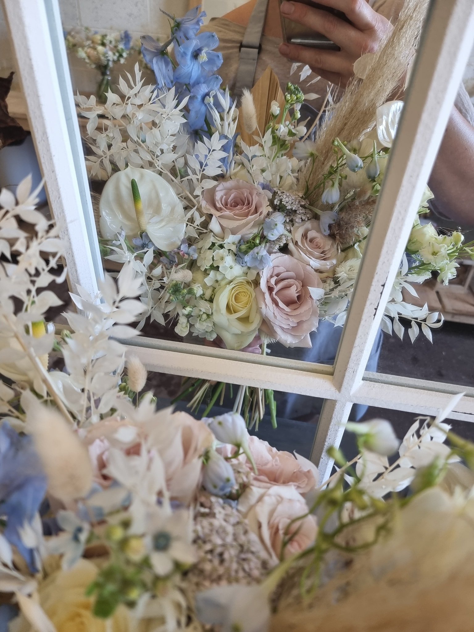 A bouquet of pastel-colored roses, blue delphiniums, and white anthuriums, reflected in a mirror, with the photographer's hand and phone visible.