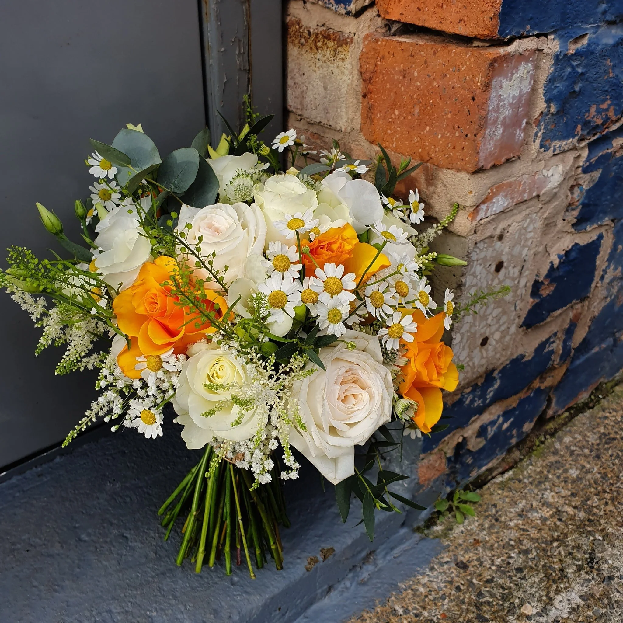 A bouquet of white, orange, and yellow flowers, including roses and daisies, placed against a brick and concrete wall.