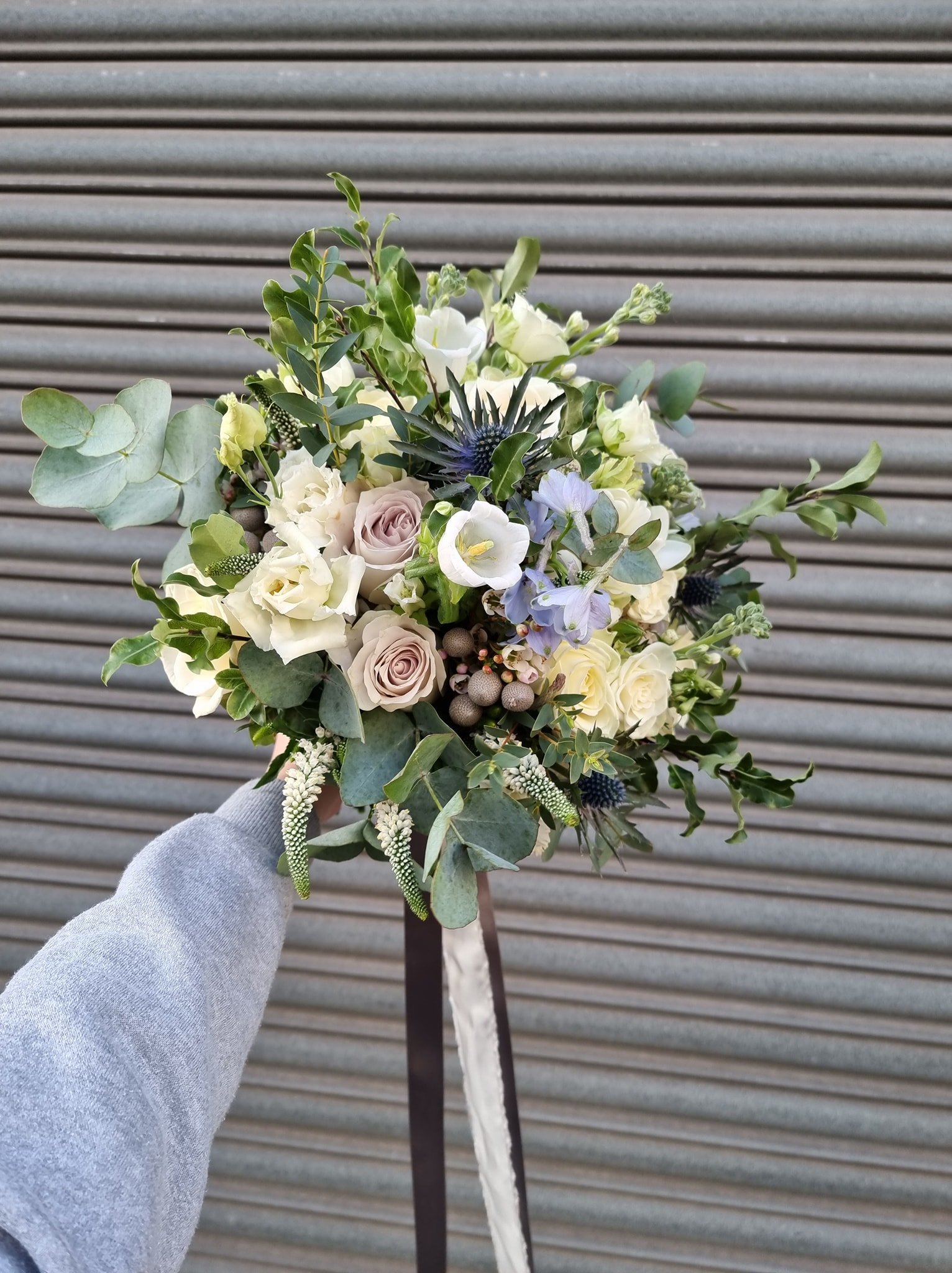 A hand holding a bouquet of white and pale pink roses, blue and white flowers, and green foliage in front of a metal shutter.