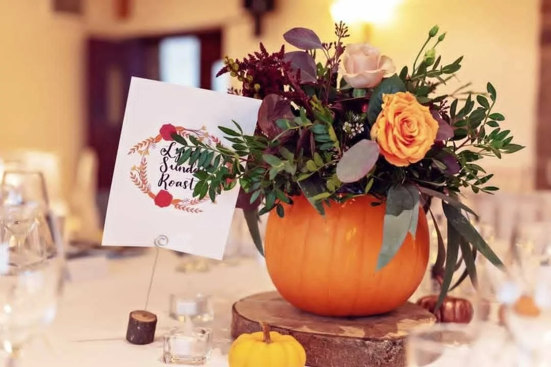 A floral centerpiece with orange and pink roses, green and purple leaves in a pumpkin-shaped vase on a wooden slab, on a table with candles and a sign that reads 'Lindy's Sunday Roast'.