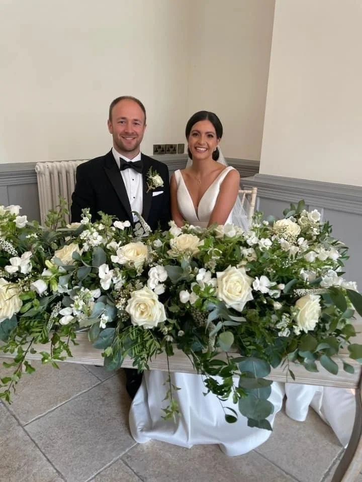 Happy bride and groom sitting behind a large floral arrangement at their wedding reception.