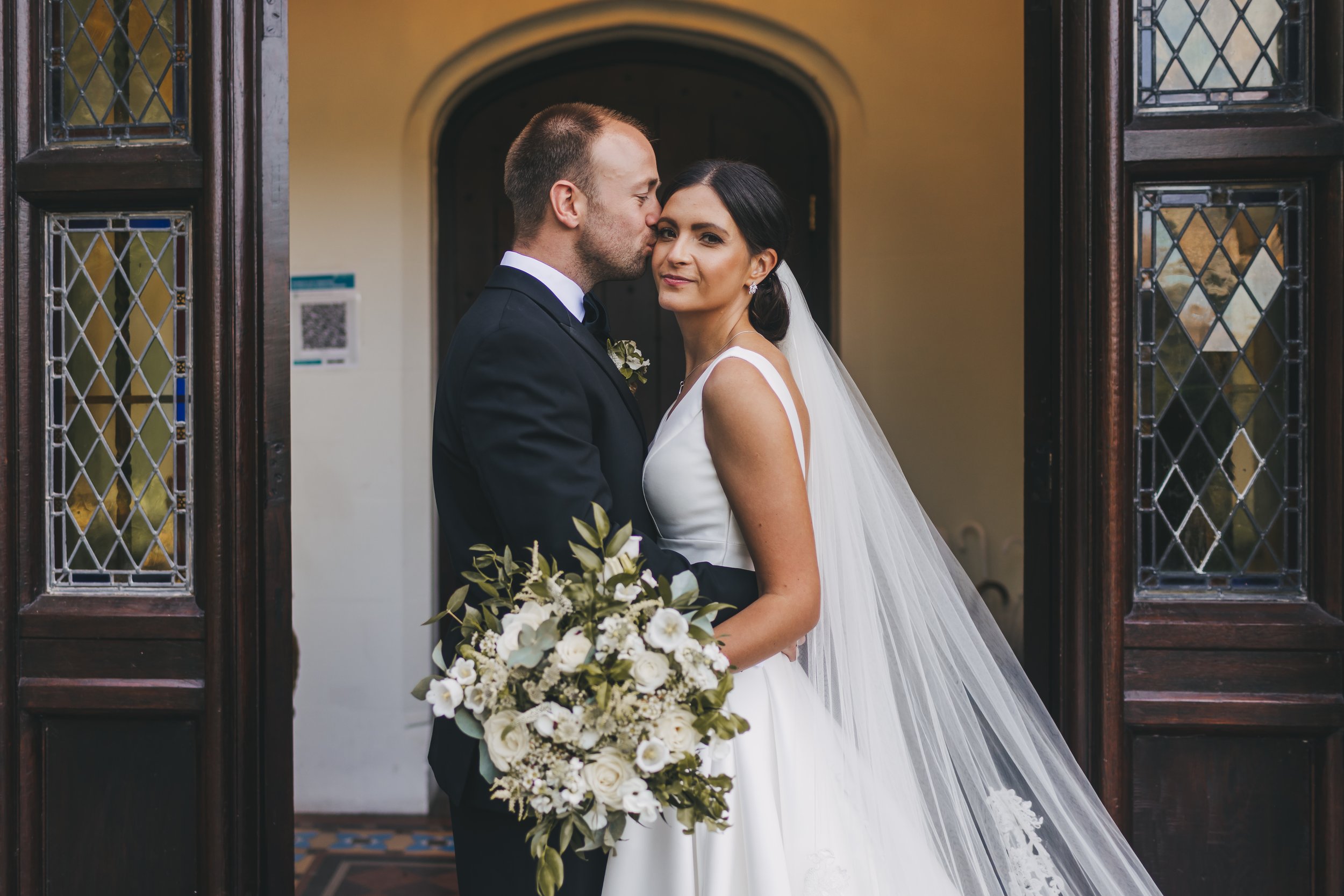 Bride and groom standing close at their wedding, with the groom kissing the bride's forehead. The bride is holding a bouquet of white flowers and wearing a white wedding gown and veil. They are in front of a wooden doorway with stained glass windows.