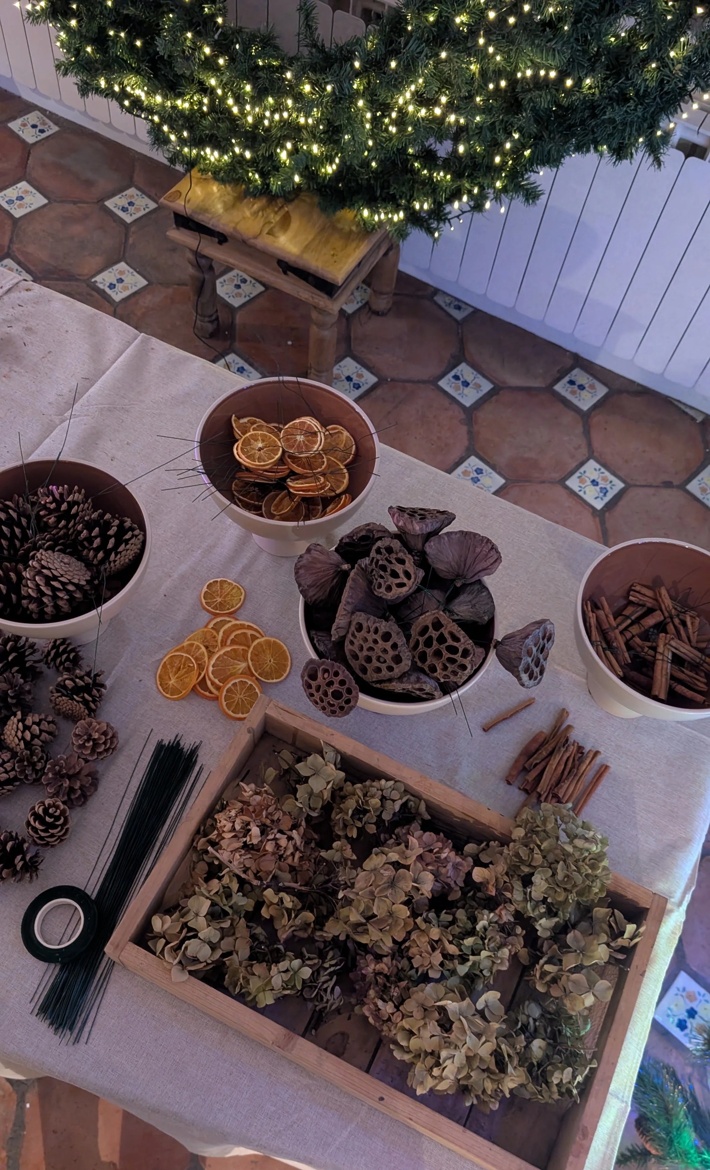 A table with dried flower arrangements, pinecones, cinnamon sticks, dried orange slices, and seed pods, with a Christmas tree decorated with lights in the background.
