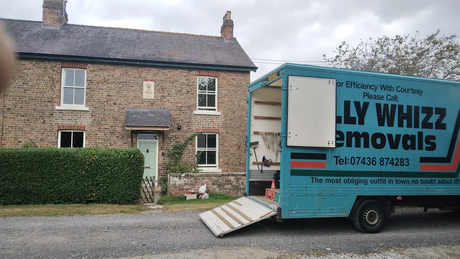 A blue moving truck parked in front of a brick house with a hedge, a small wooden gate, and a garden with animal figurines. The truck has an open side with tools and a ramp.