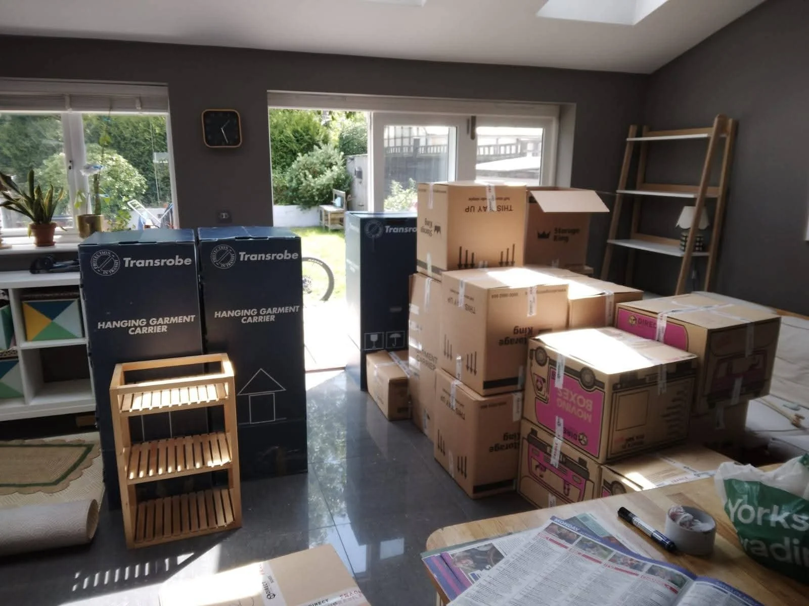 Living room filled with unpacked moving boxes, including black Transrobe boxes labeled "Hanging Garment Carrier" and various other boxes stacked near a window and door, with a wooden shelf and a table with newspapers and markers.
