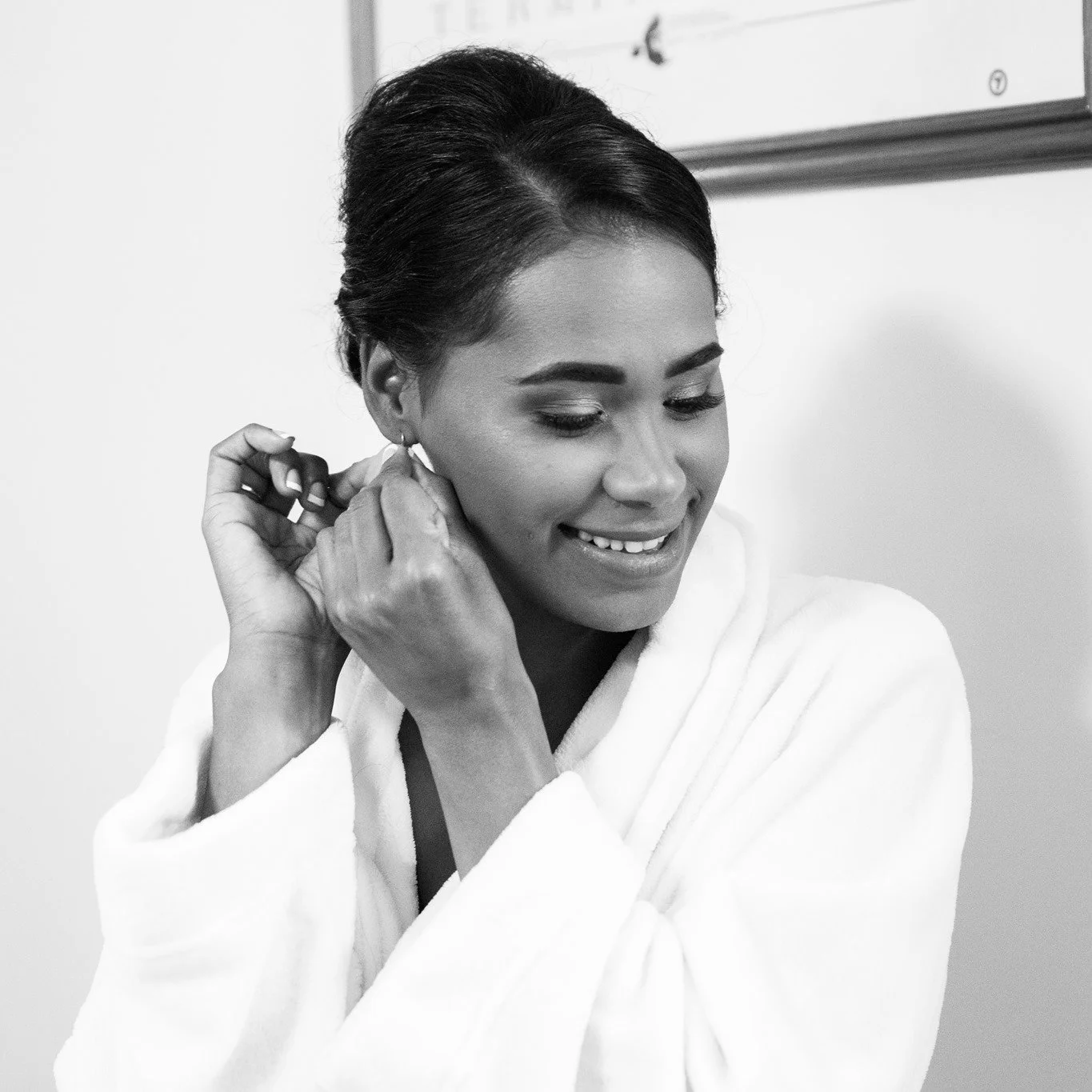 A woman with styled dark hair and makeup, wearing a white bathrobe, smiling as she puts on earrings.