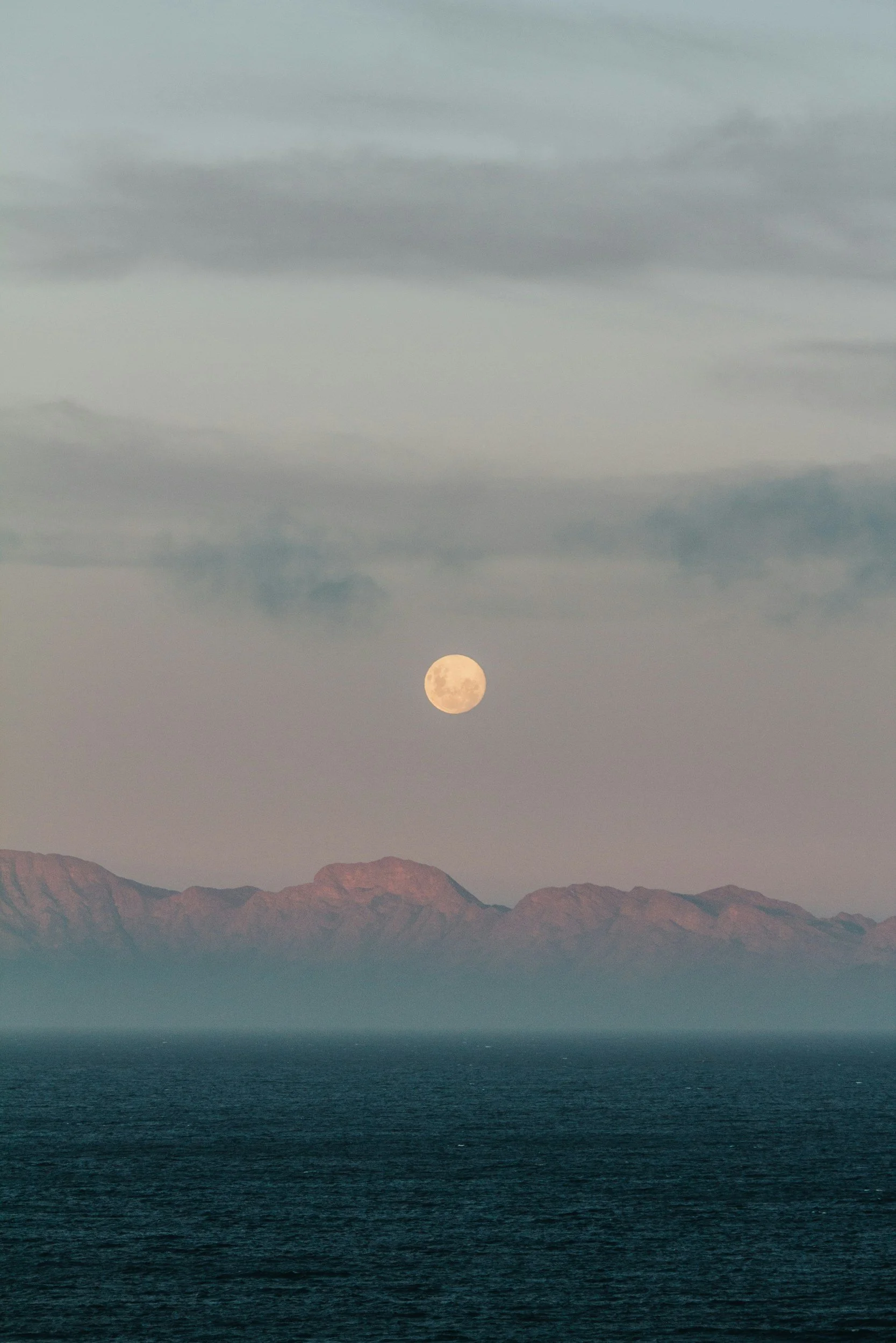 Full moon rising over a mountain range with an ocean in the foreground and clouds in the sky.