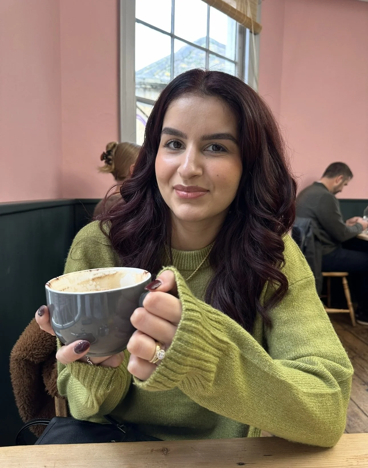 A young woman with dark wavy hair, wearing a green sweater, holding a coffee mug, sitting inside a cafe with pink walls and a large window behind her.