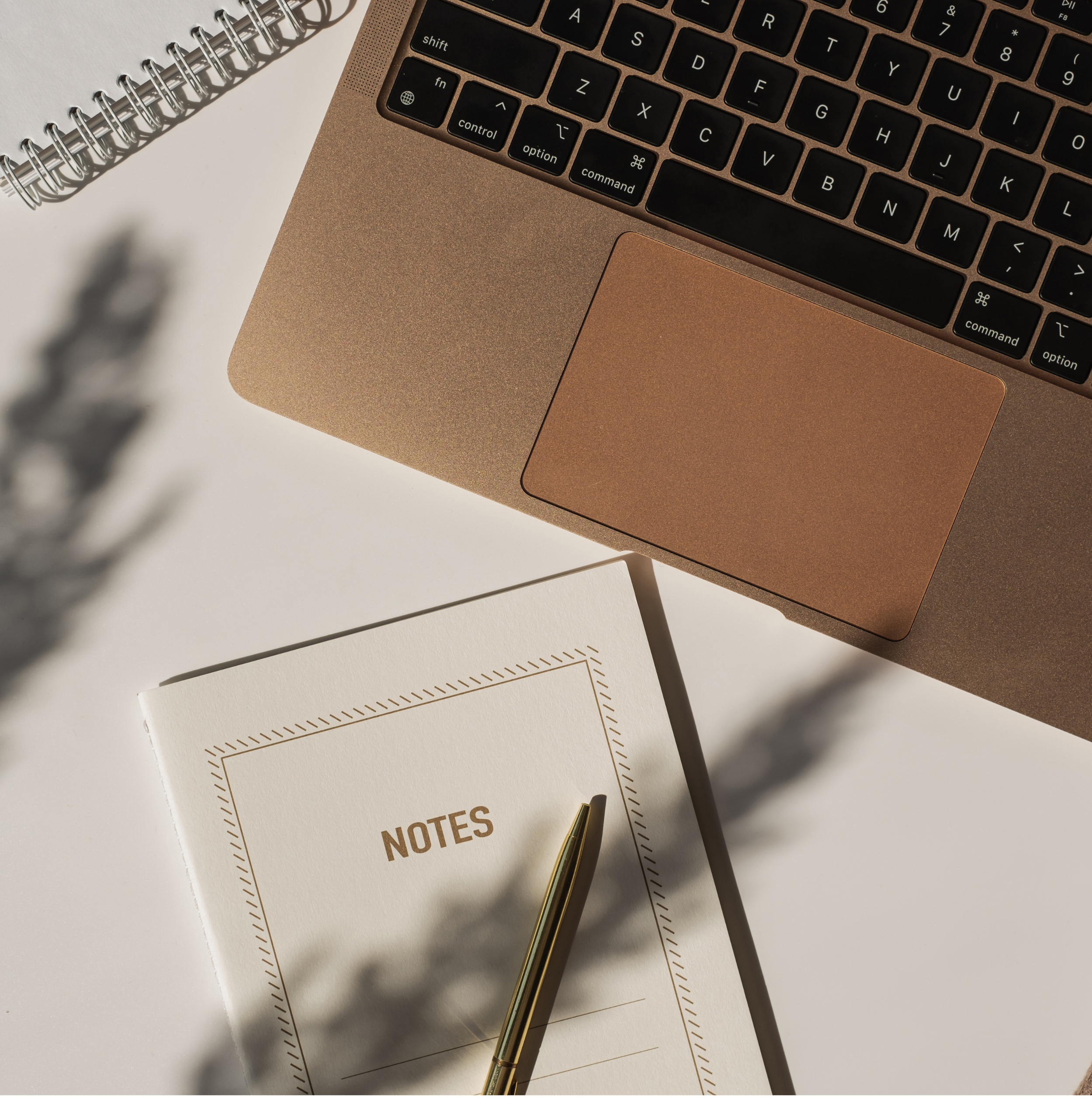 Close-up of a workspace showing a gold-colored laptop, a white notebook with "NOTES" written on it, and a gold pen on a white surface with shadows