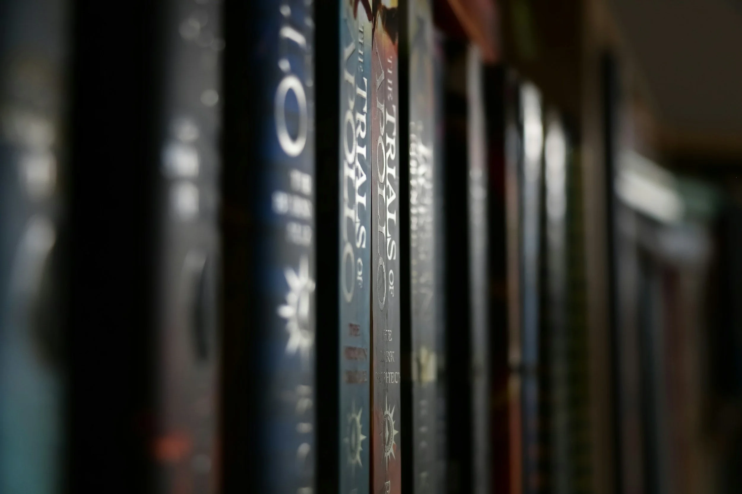Close-up of books on a shelf, focusing on their spines with titles partially visible, in low lighting.