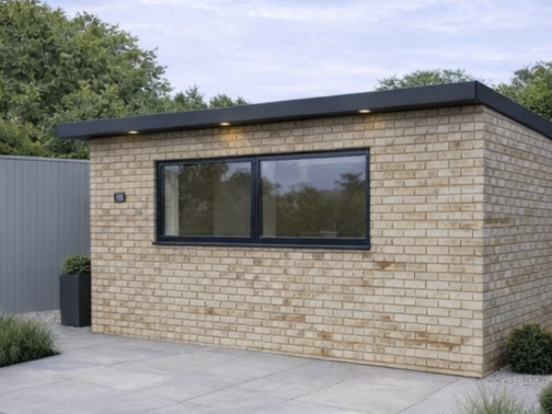A completed Steel Frame Garden Room with a large window, black trim, and a flat roof, surrounded by a paved patio and greenery.