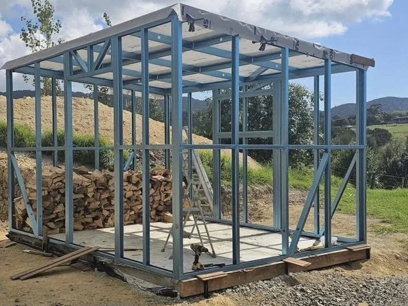 Construction of a small metal frame structure, with wooden logs stacked in the background, a ladder, and construction tools outside on a dirt ground in an outdoor rural setting with hills and trees in the distance.