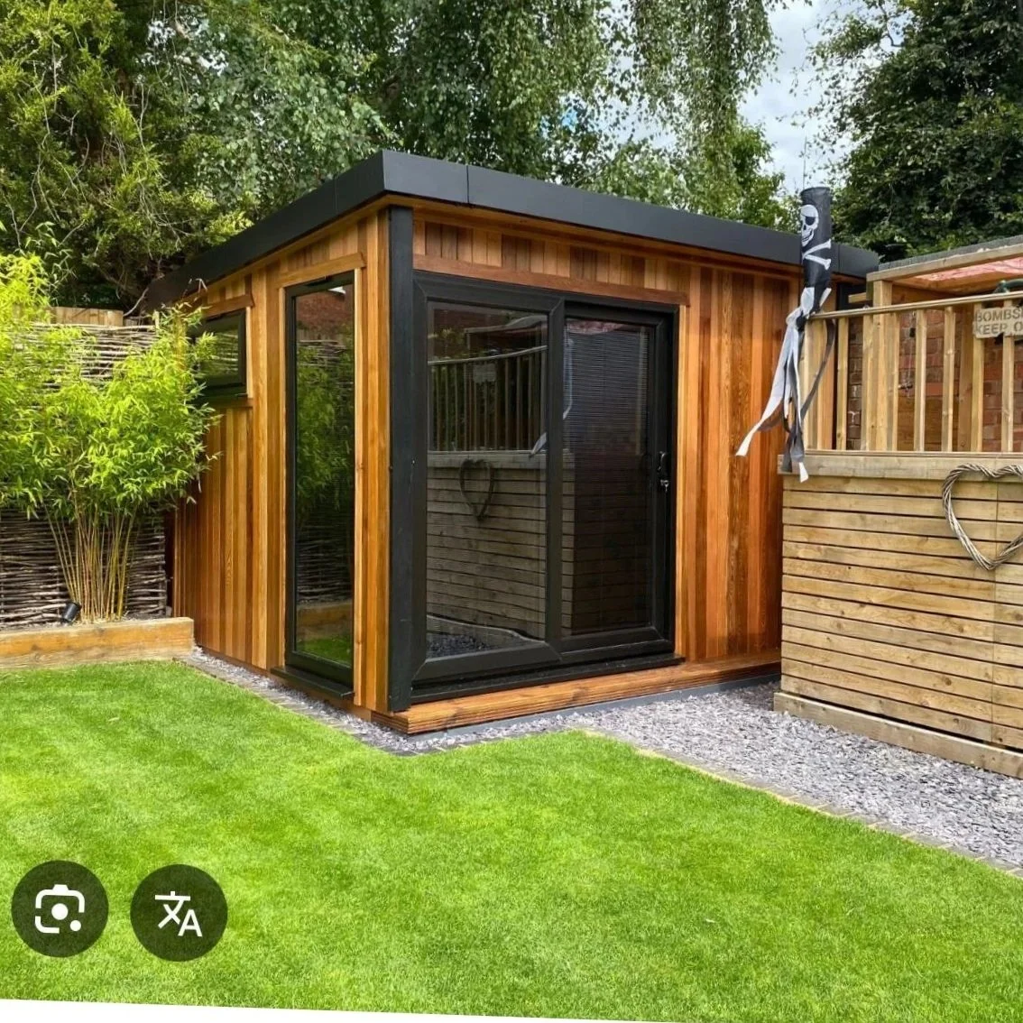 A small wooden garden rpom with black trim and a glass door, surrounded by a green lawn and trees.