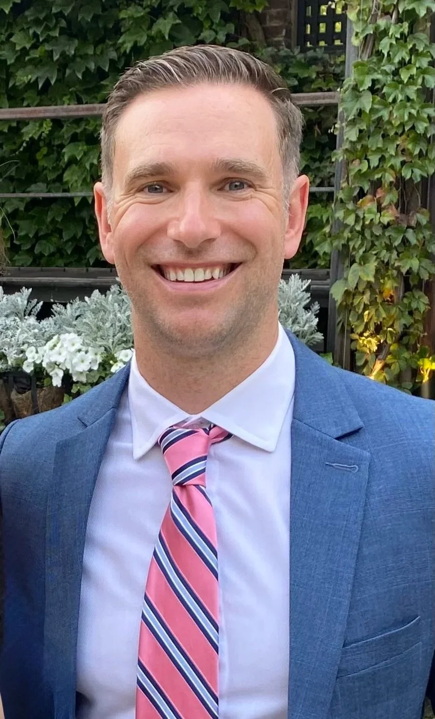A man smiling, wearing a blue suit, white shirt, and a pink, blue, and white striped tie, standing outdoors with green foliage and white flowers in the background.