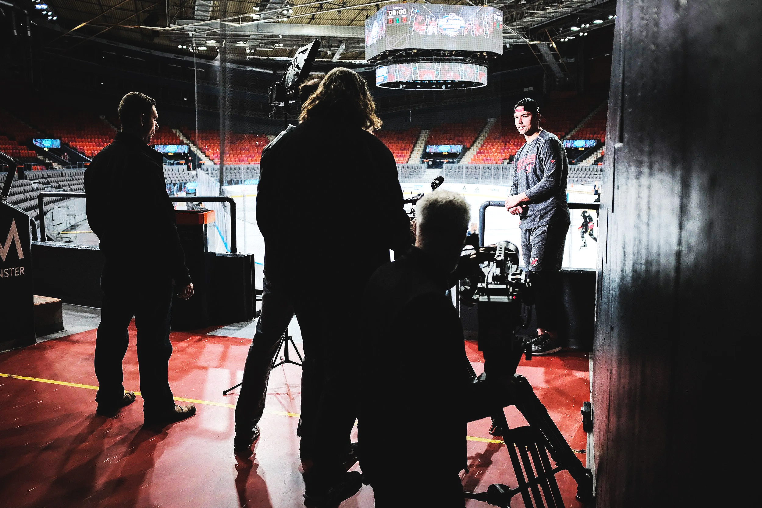 A person being interviewed in an arena with an ice hockey rink in the background, along with camera operators and crew recording the interview.