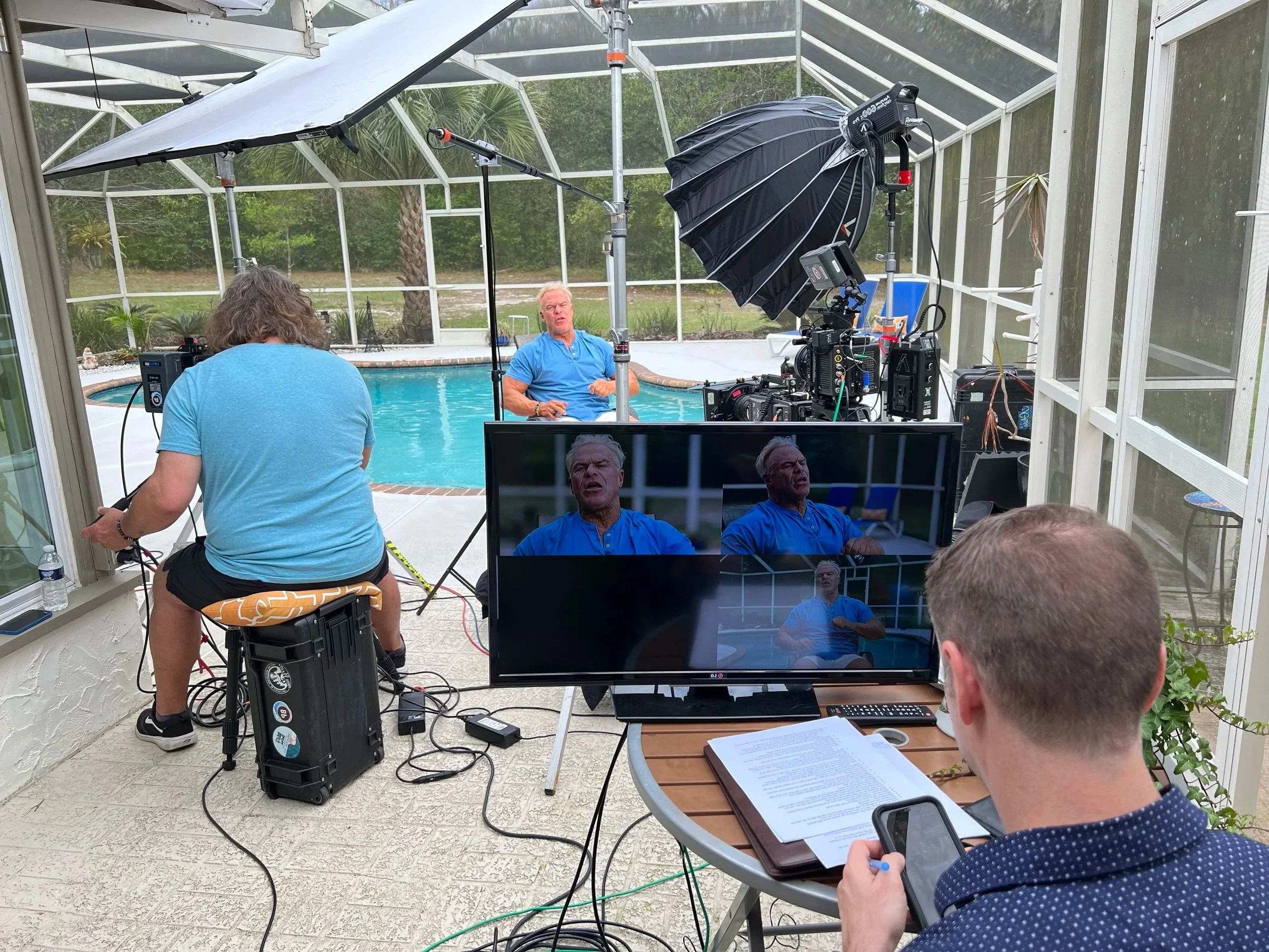 A behind-the-scenes view of a video interview setup by a swimming pool, with multiple cameras, lighting, monitors, and crew members preparing for the recording.