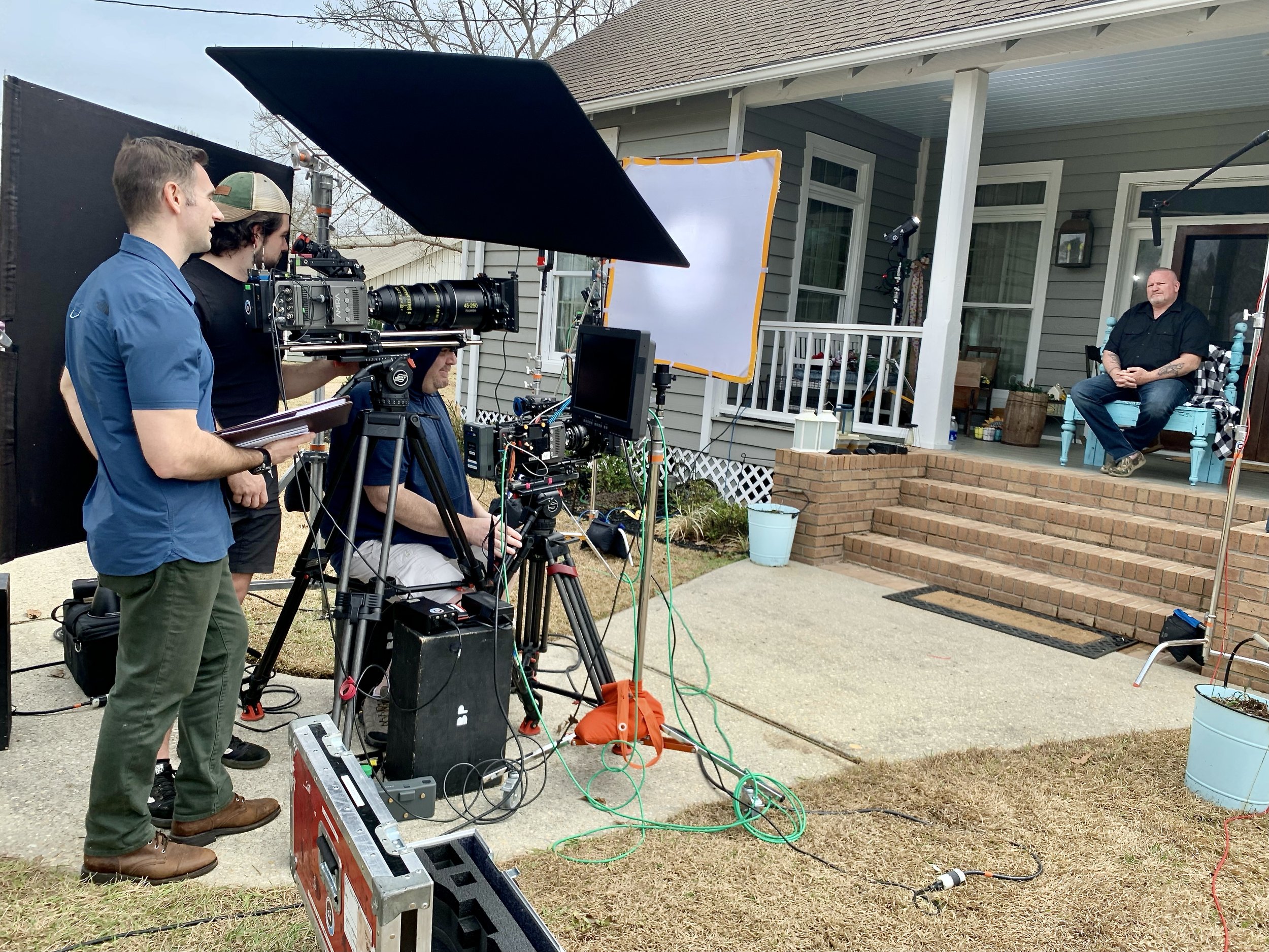 Filming crew sets up equipment outside a house for an interview or scene on the front porch, with a man sitting on a blue bench in the background.