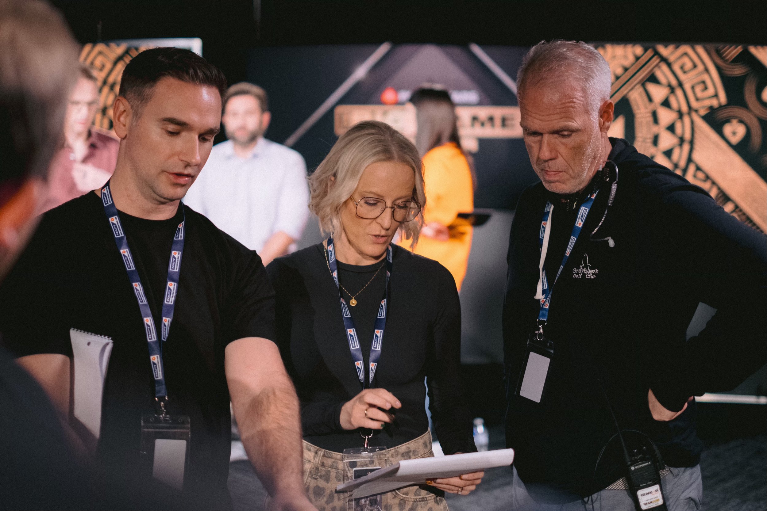 Group of people working together at a professional event, wearing lanyards and discussing documents, with a backdrop featuring banners and decorative patterns.