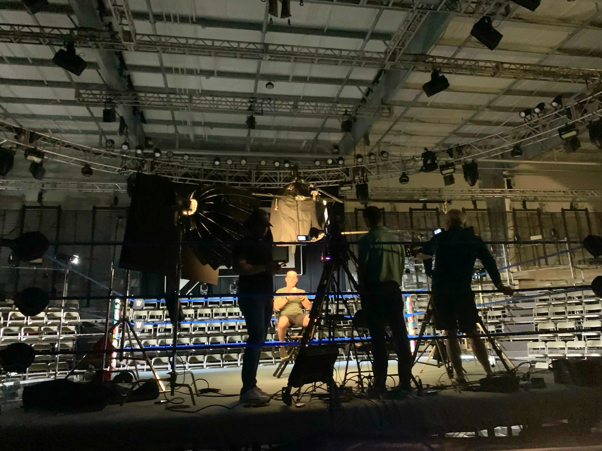 Interior of a photography or video shoot setup with crew members preparing in front of a seated person in a partially lit arena or event space.