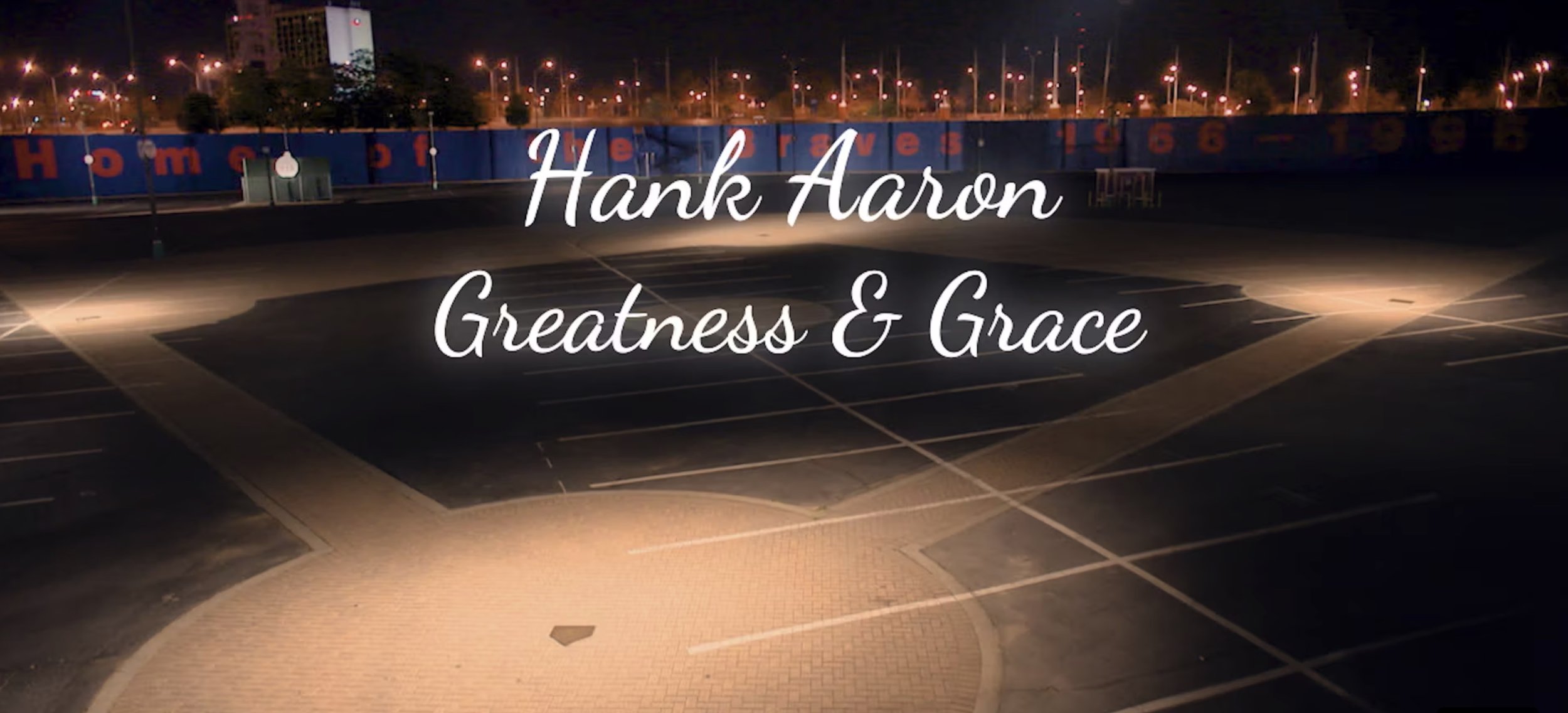 Empty parking lot at night with a blue wall in the background displaying numbers and the word 'Homes' in red, and streetlights illuminating the area. Text overlay reads 'Hank Aaron Greatness & Grace' in white cursive font.