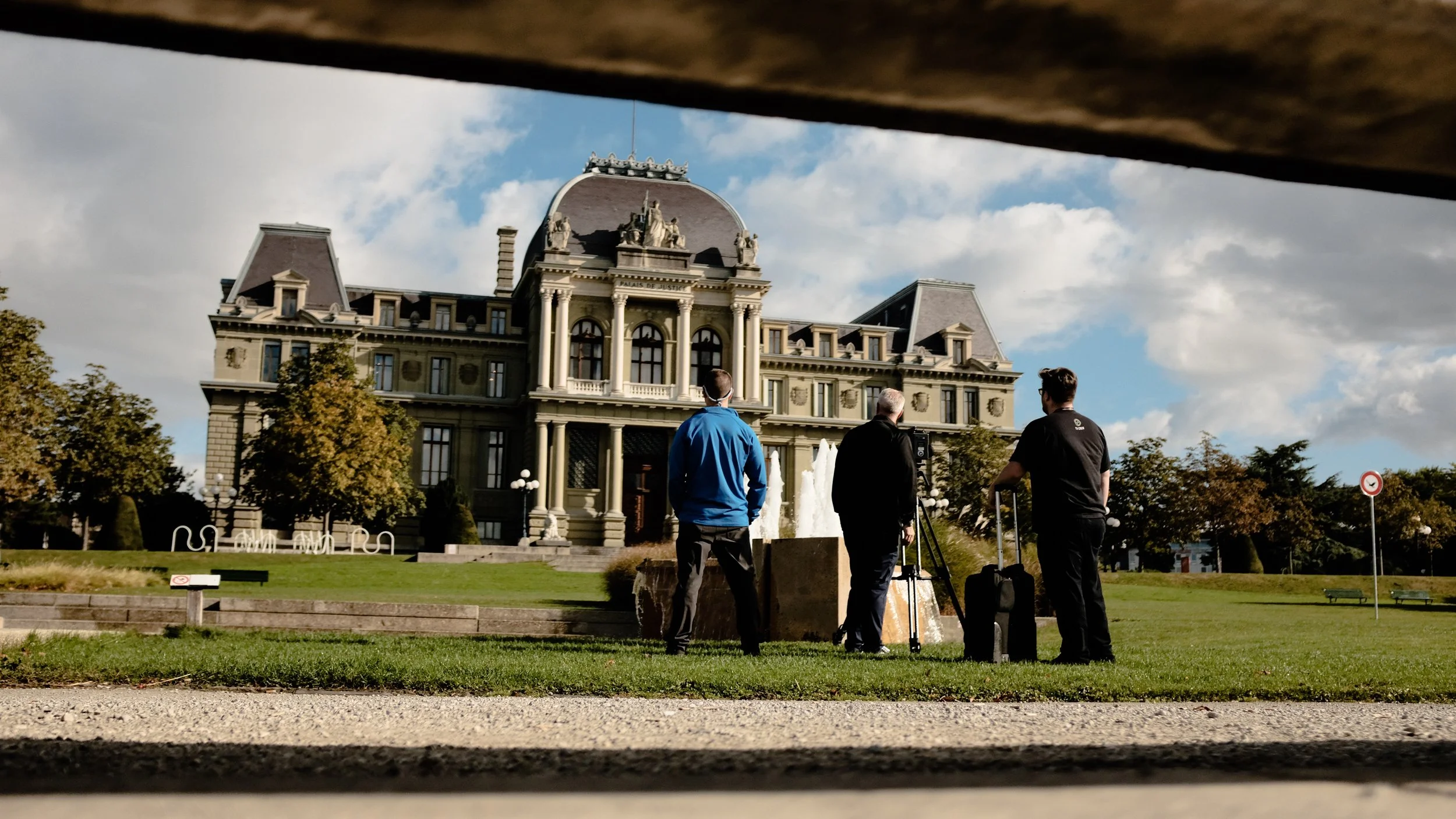 A historical building with ornate architecture, including columns and statues, seen from a low angle through a gap. In front of the building, three men are standing on the grass, with luggage and equipment, facing the building, under a partly cloudy sky.
