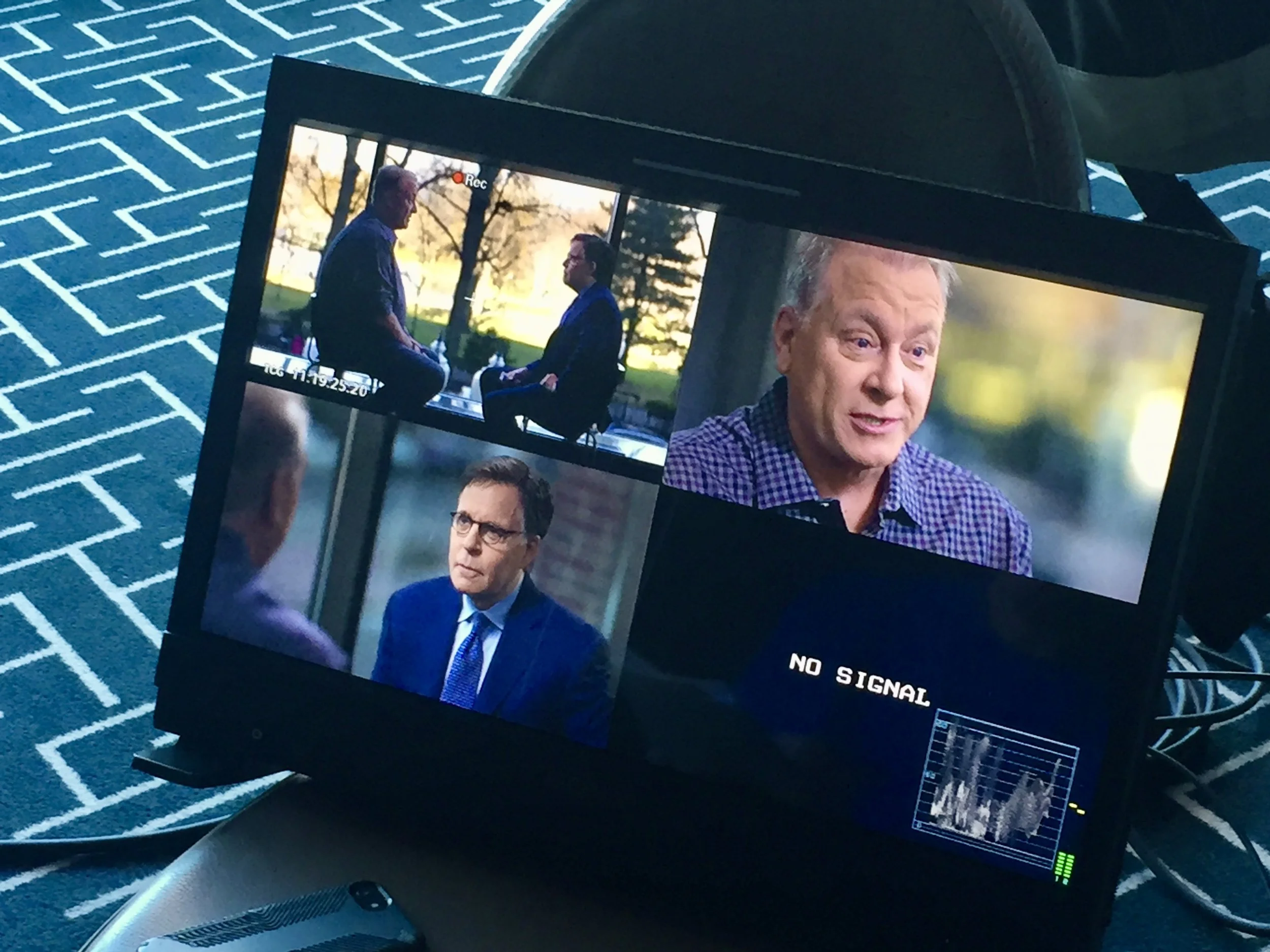 Video camera monitor shows scenes from an interview, including two men conversing outdoors against a park background, and a close-up of a man speaking, with 'NO SIGNAL' message displayed on part of the monitor.