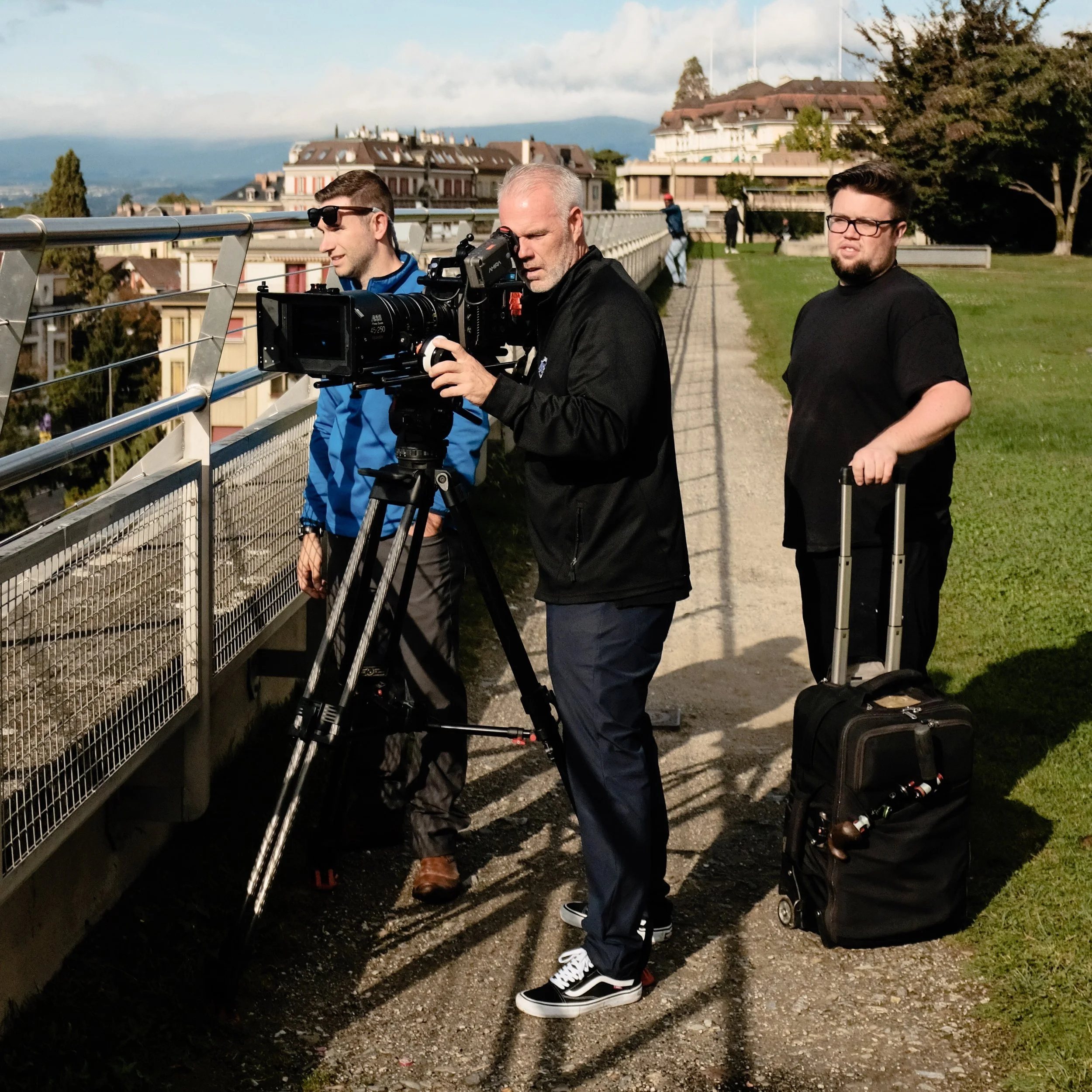 Three men filming with a professional camera on a tripod, standing outdoors on a sunny day near a cityscape and greenery.