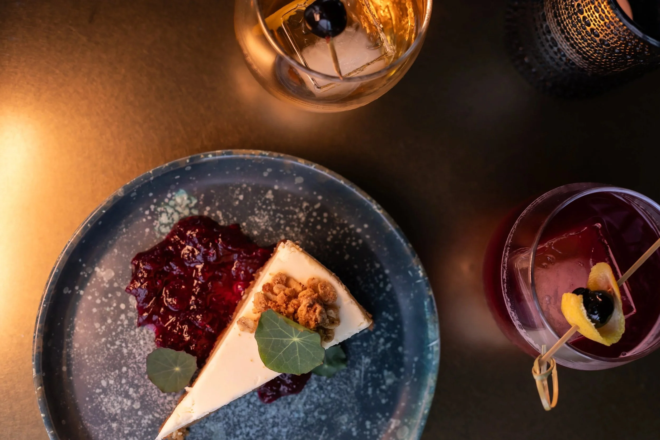 A slice of cheesecake on a dark speckled plate garnished with a green leaf, served with berry compote. Two cocktails, one with ice and a black cherry, and another with lemon and a maraschino cherry, are on a dark table, illuminated by warm lighting.