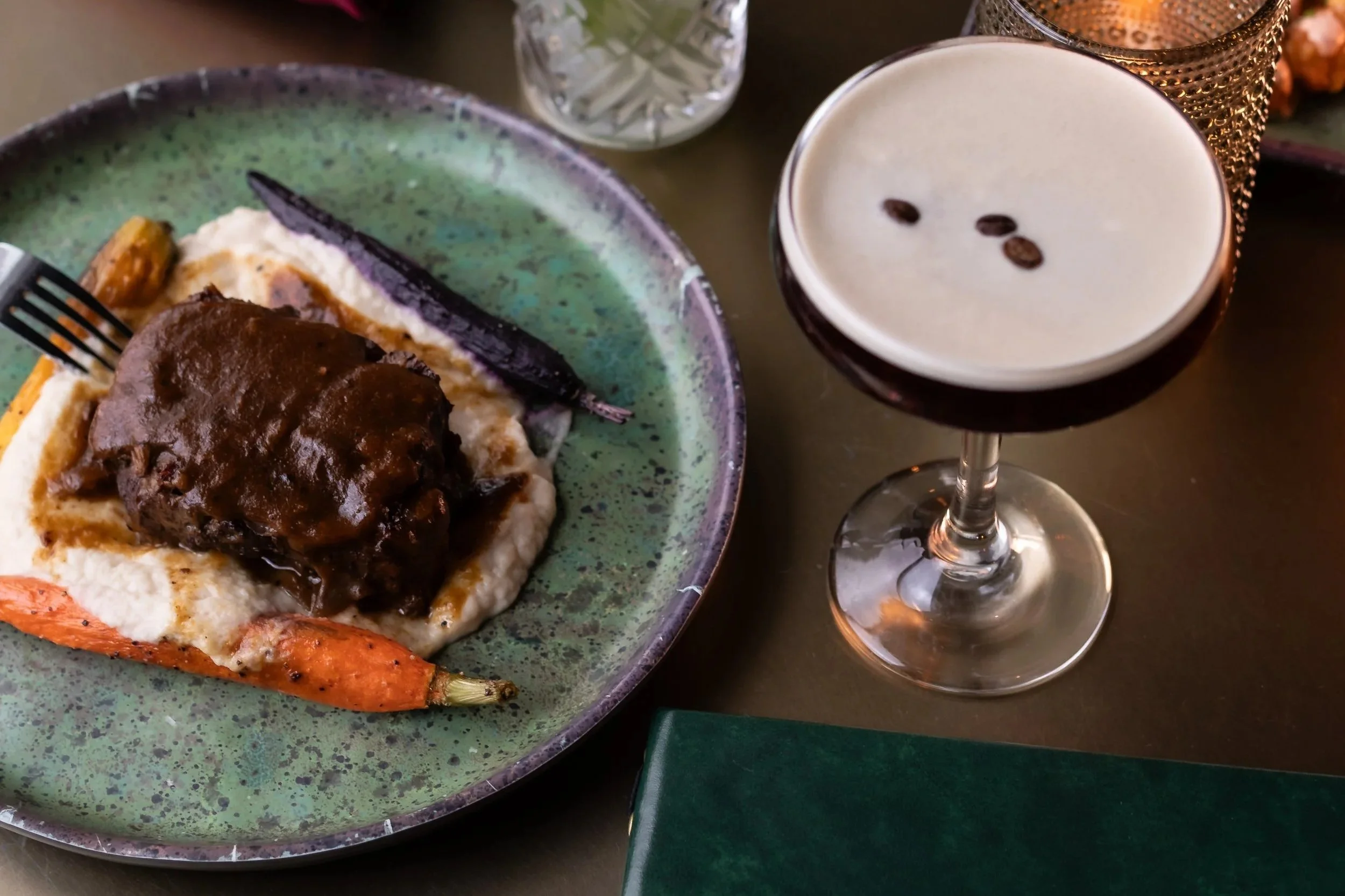 Plate of mashed potatoes topped with beef gravy, roasted carrots, and a slice of purple onion, with an espresso martini with three coffee beans on top.