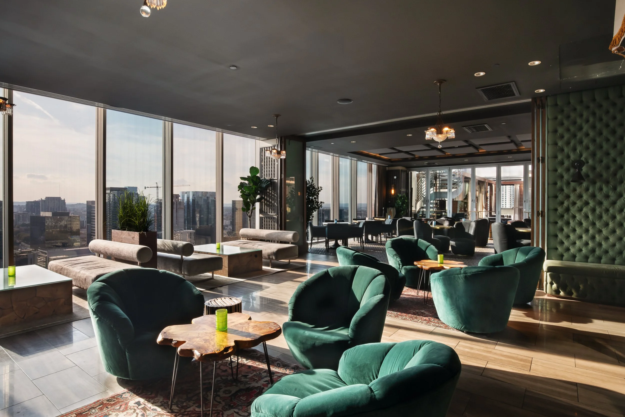 Modern high-rise lounge with green velvet armchairs, wooden tables, large floor-to-ceiling windows, city view, and stylish lighting fixtures.