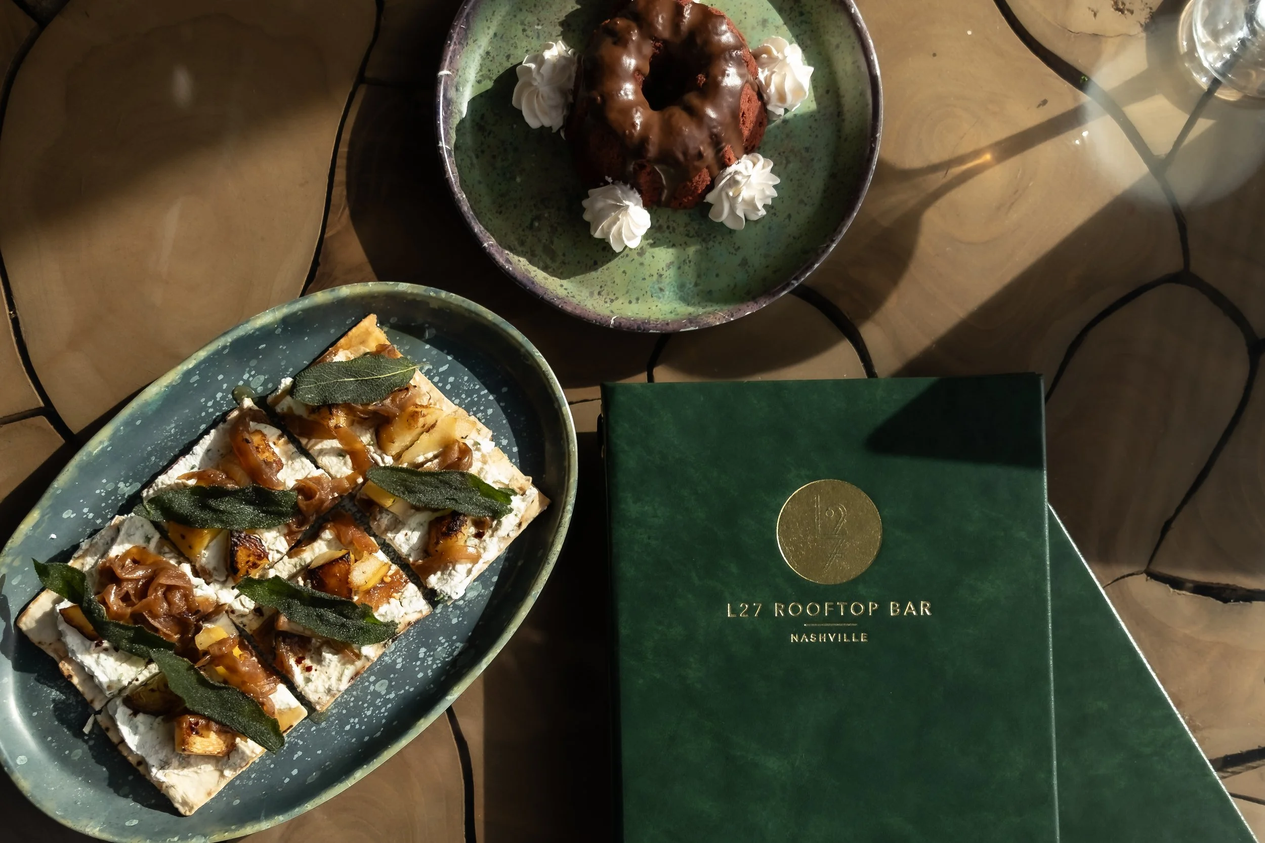 Two appetizers on a table: a rectangular plate with caramelized nuts and herbs on a cheese or frosted cracker, a round chocolate cake with caramel and dollops of whipped cream, and a green menu labeled 'L27 Rooftop Bar Nashville'.