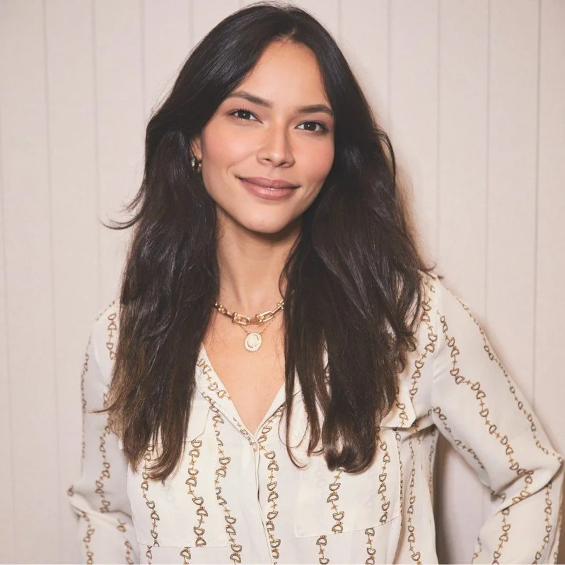 A woman with long dark hair, wearing a white blouse with golden embroidery, gold jewelry, and a necklace with a pendant, standing against a white wooden background.