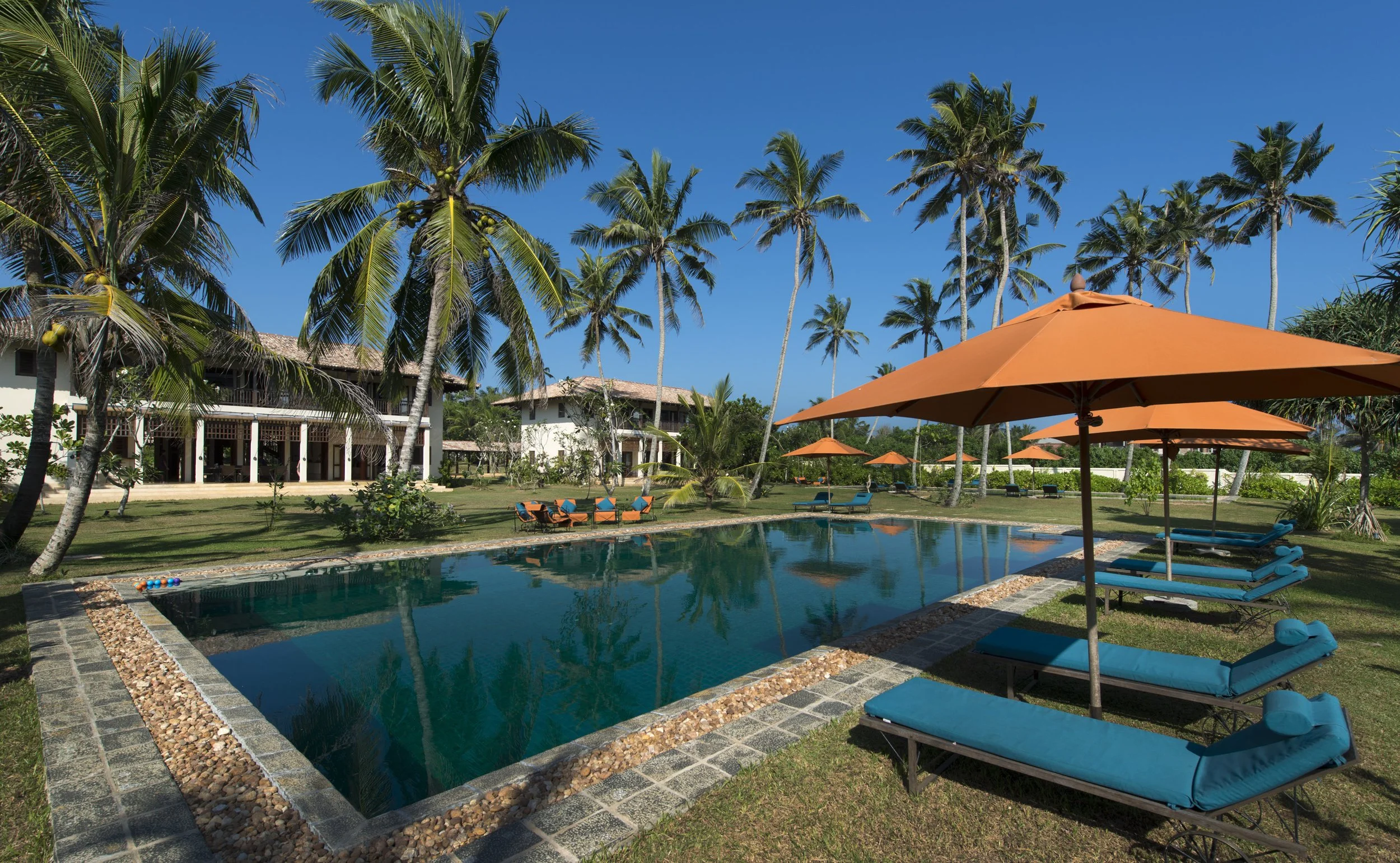 A tropical resort with a swimming pool, surrounded by palm trees, with orange umbrellas and blue lounge chairs, and a building with a balcony in the background under a clear blue sky.